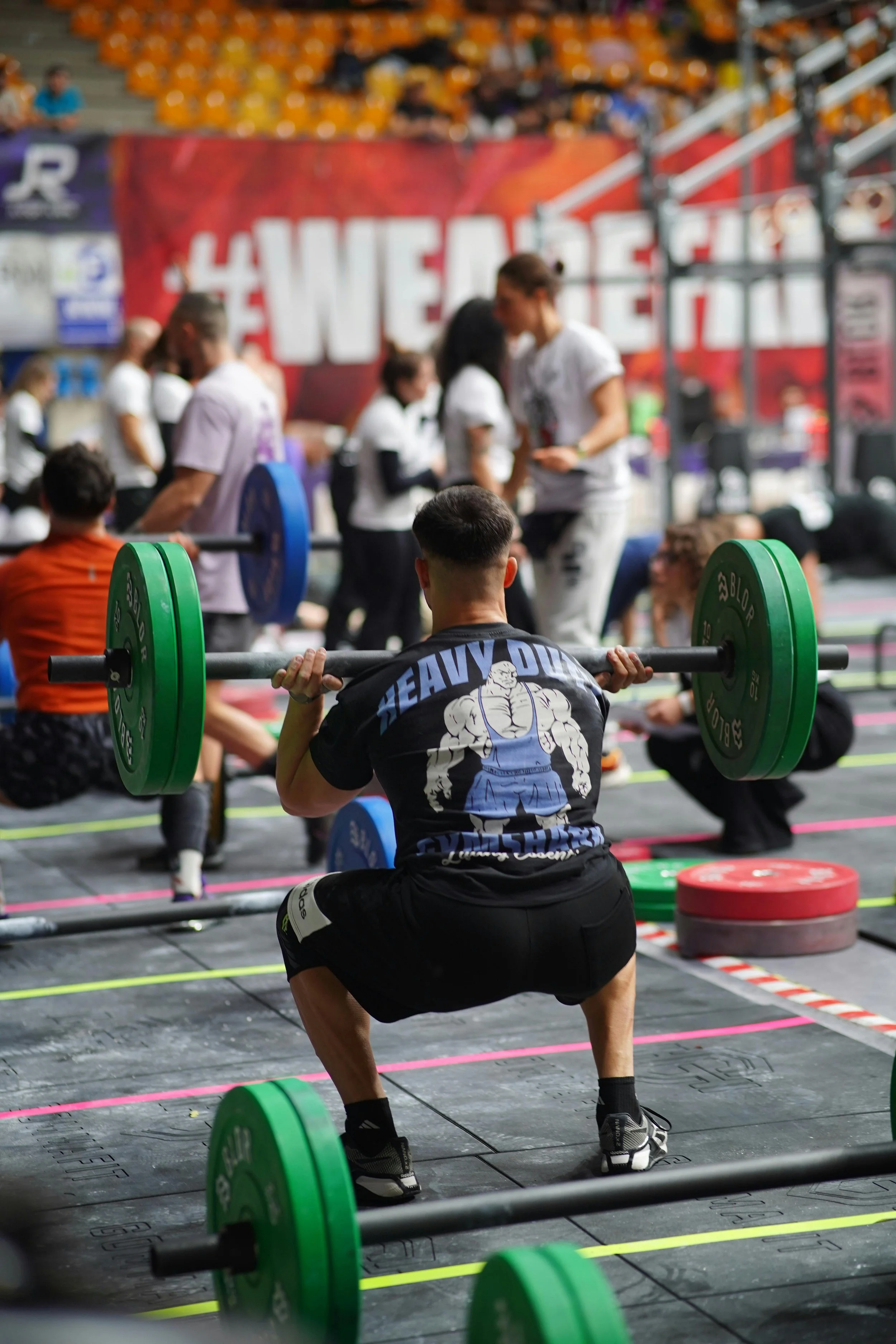 A man performing a squat exercise with a barbell loaded with green and blue weights at a powerlifting competition.