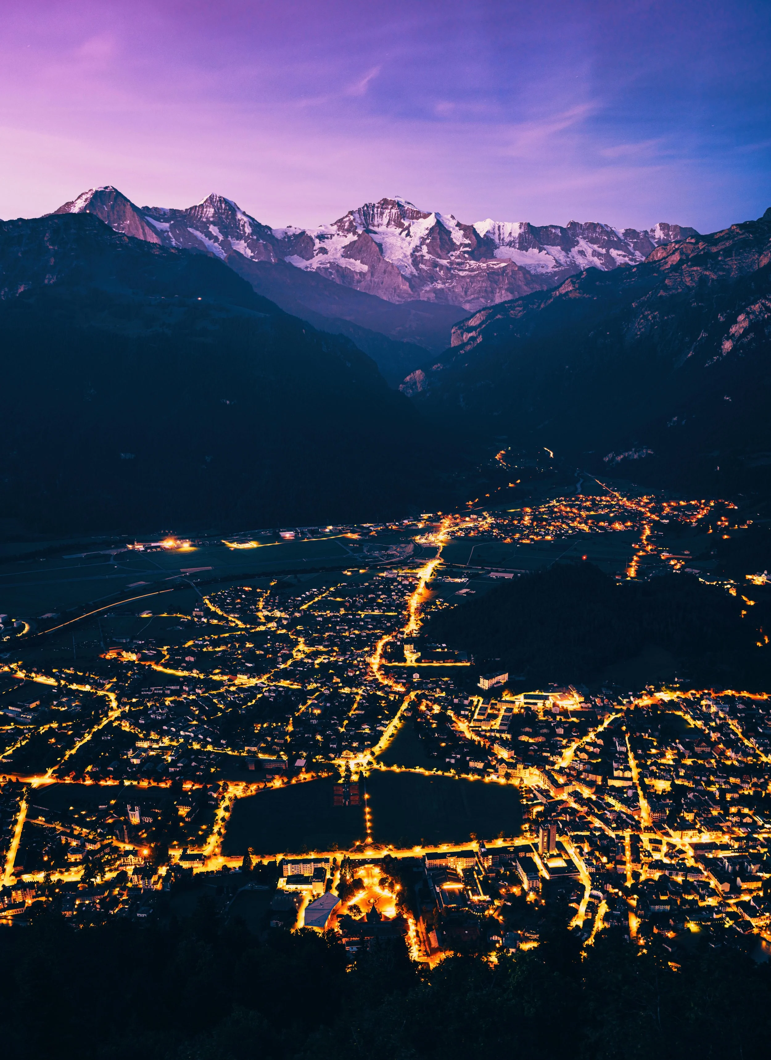 Night view of a illuminated town in a valley with mountains in the background and a purple sky.