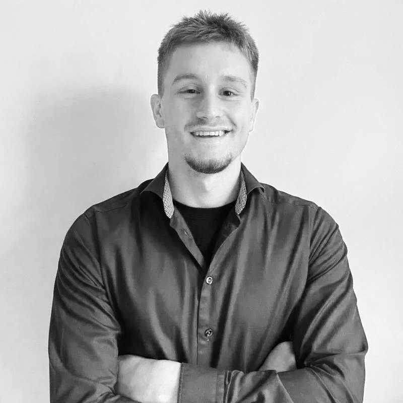 Black and white portrait of a young man with short hair, smiling, wearing a button-up shirt with arms crossed, standing against a plain wall.