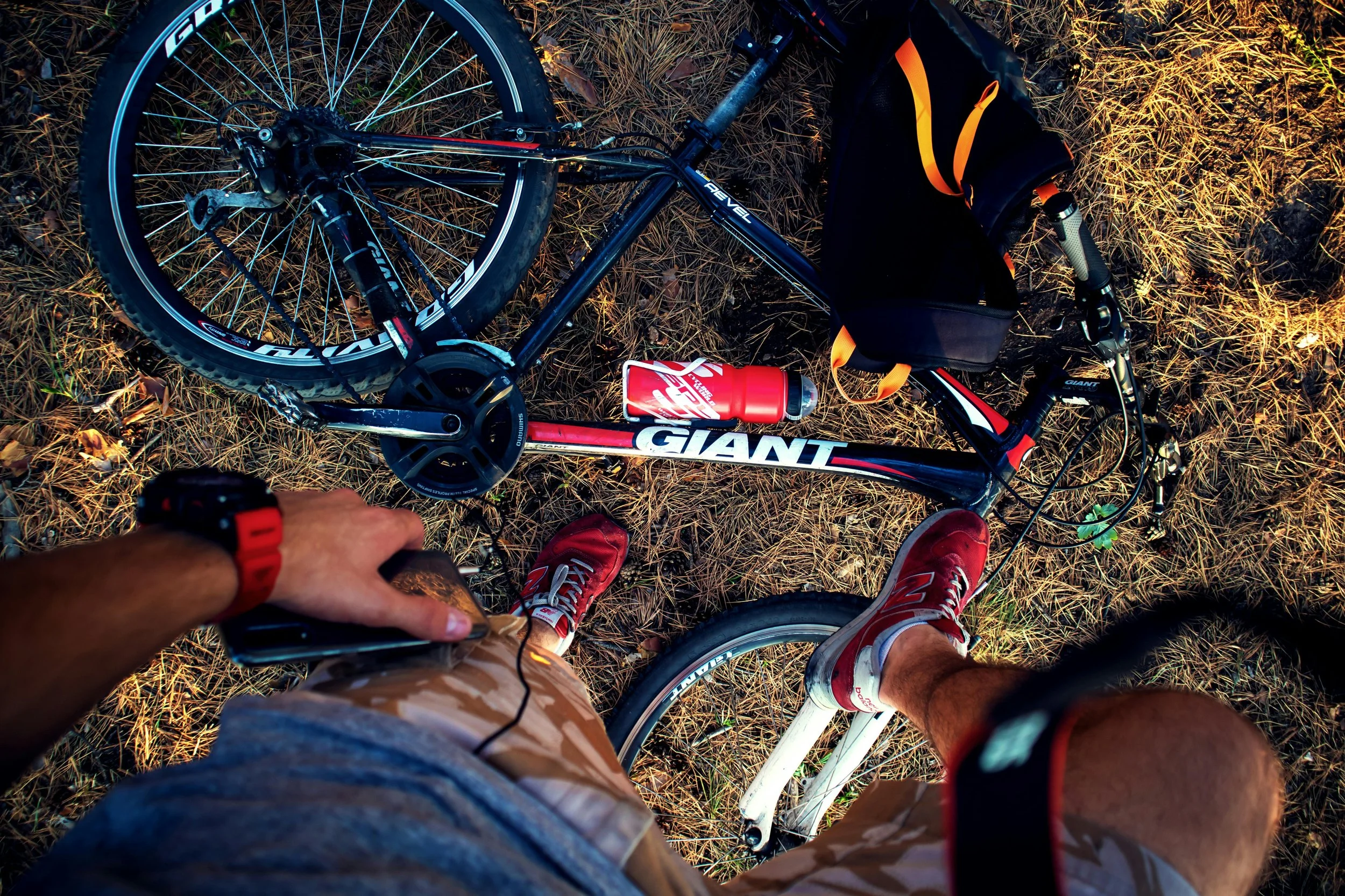Top-down view of a person standing beside their black and red Giant mountain bike on a grassy surface, with a black backpack, a red water bottle, and the person's legs and feet visible, wearing red and gray sneakers and camouflage shorts.