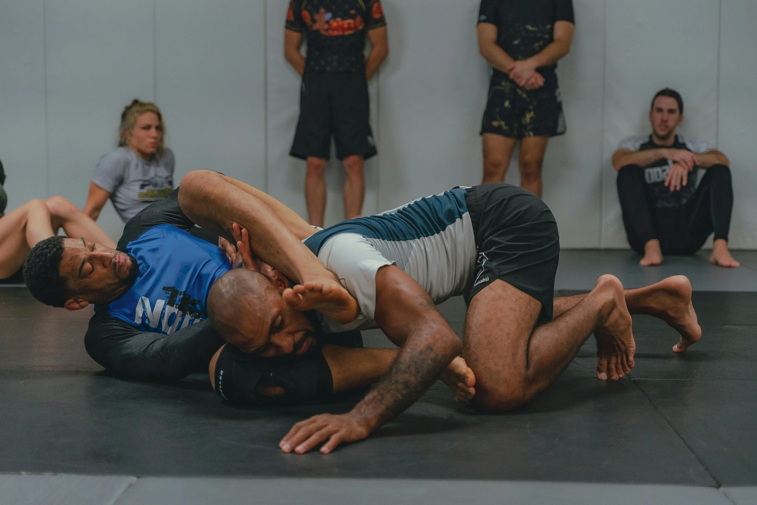 Two men competing in Brazilian Jiu-Jitsu grappling, with one man on top attempting a chokehold while the other defends, and several people observing in the background.