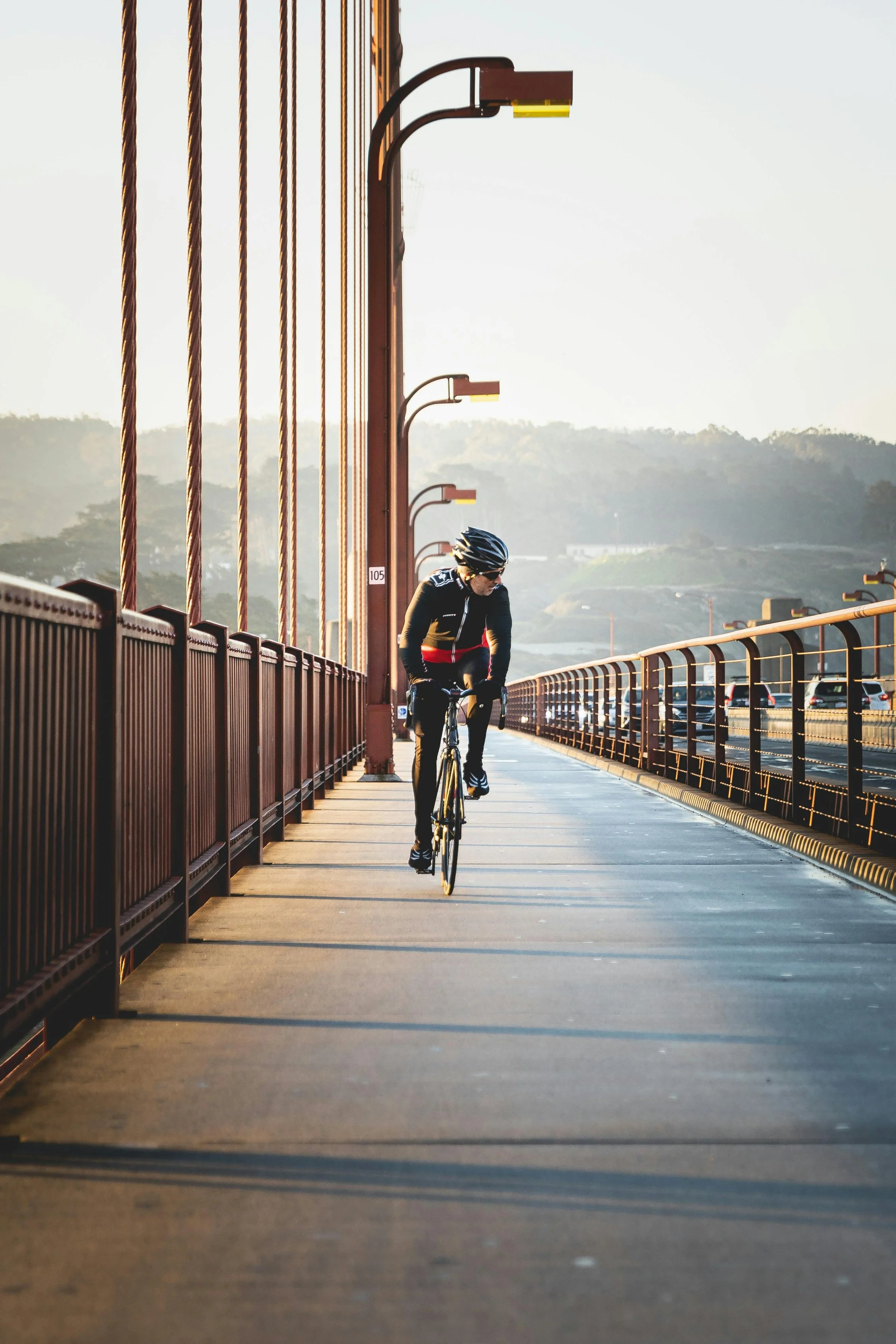 A person riding a bike on a bridge during sunrise or sunset, wearing a helmet and athletic clothing, with a scenic background of hills and traffic on the side.