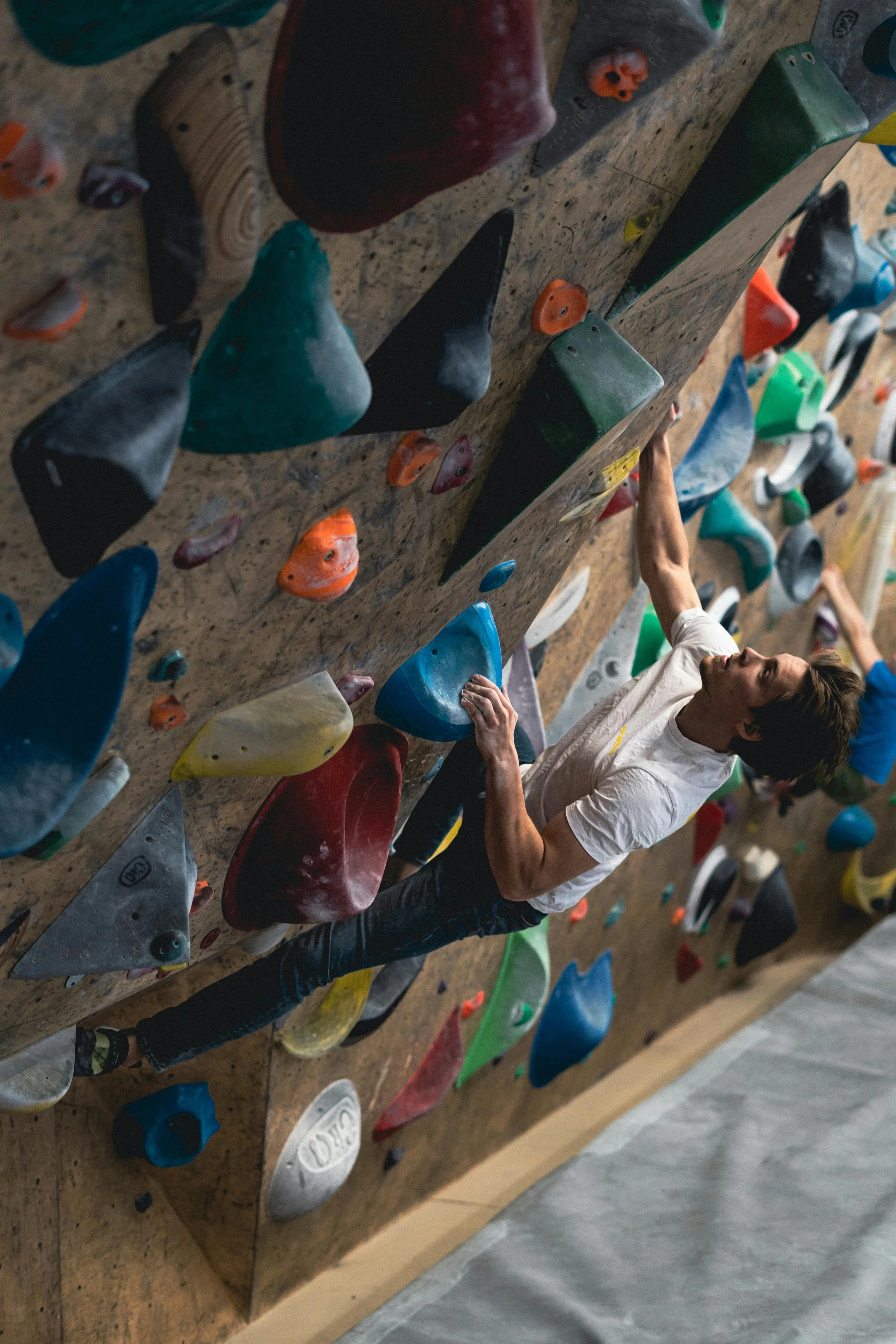 A young man with brown hair holds onto colorful climbing holds on an indoor rock climbing wall, reaching for the next hold.