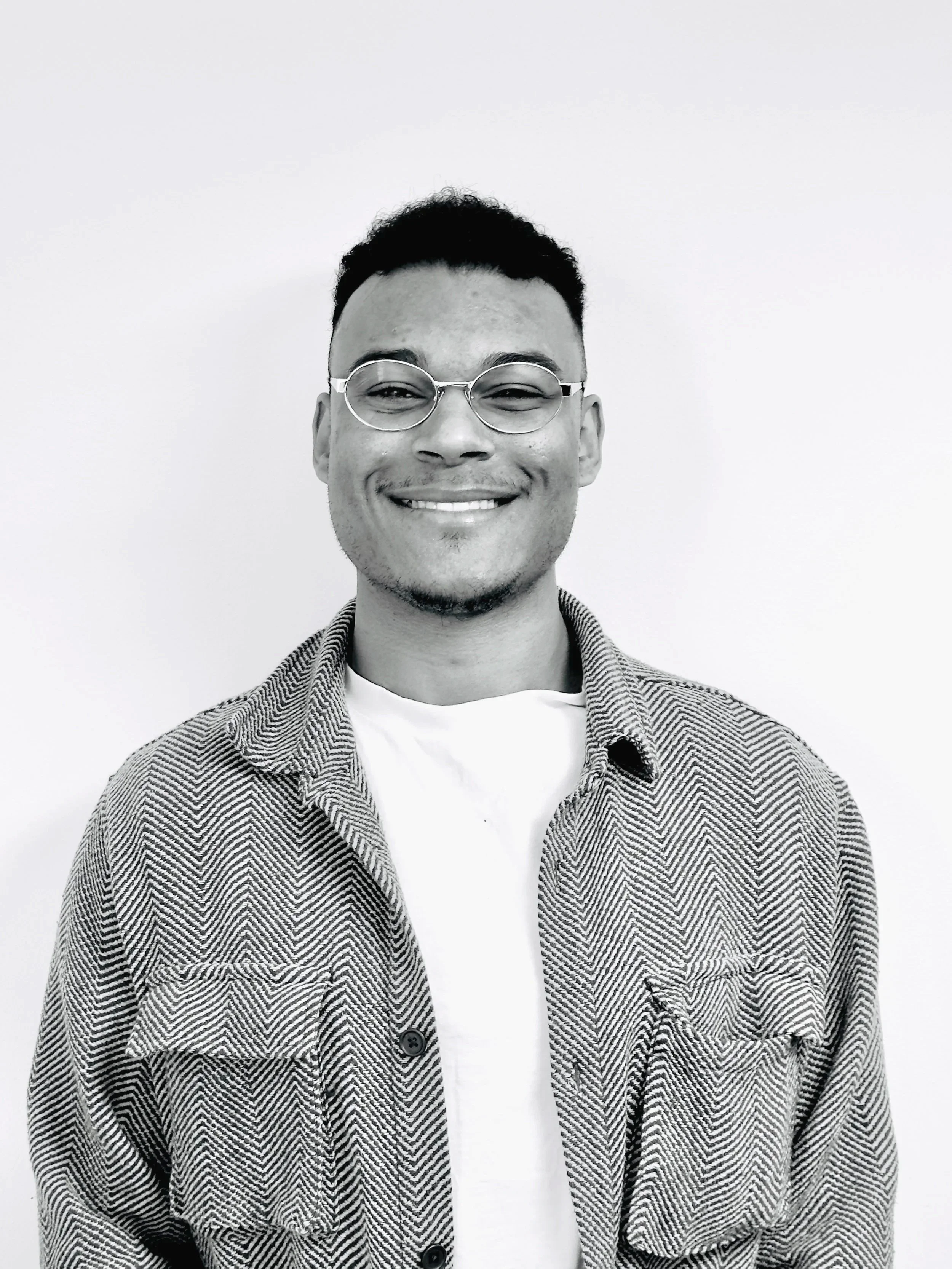 A smiling man with glasses wearing a patterned jacket over a white shirt, standing against a plain white background.