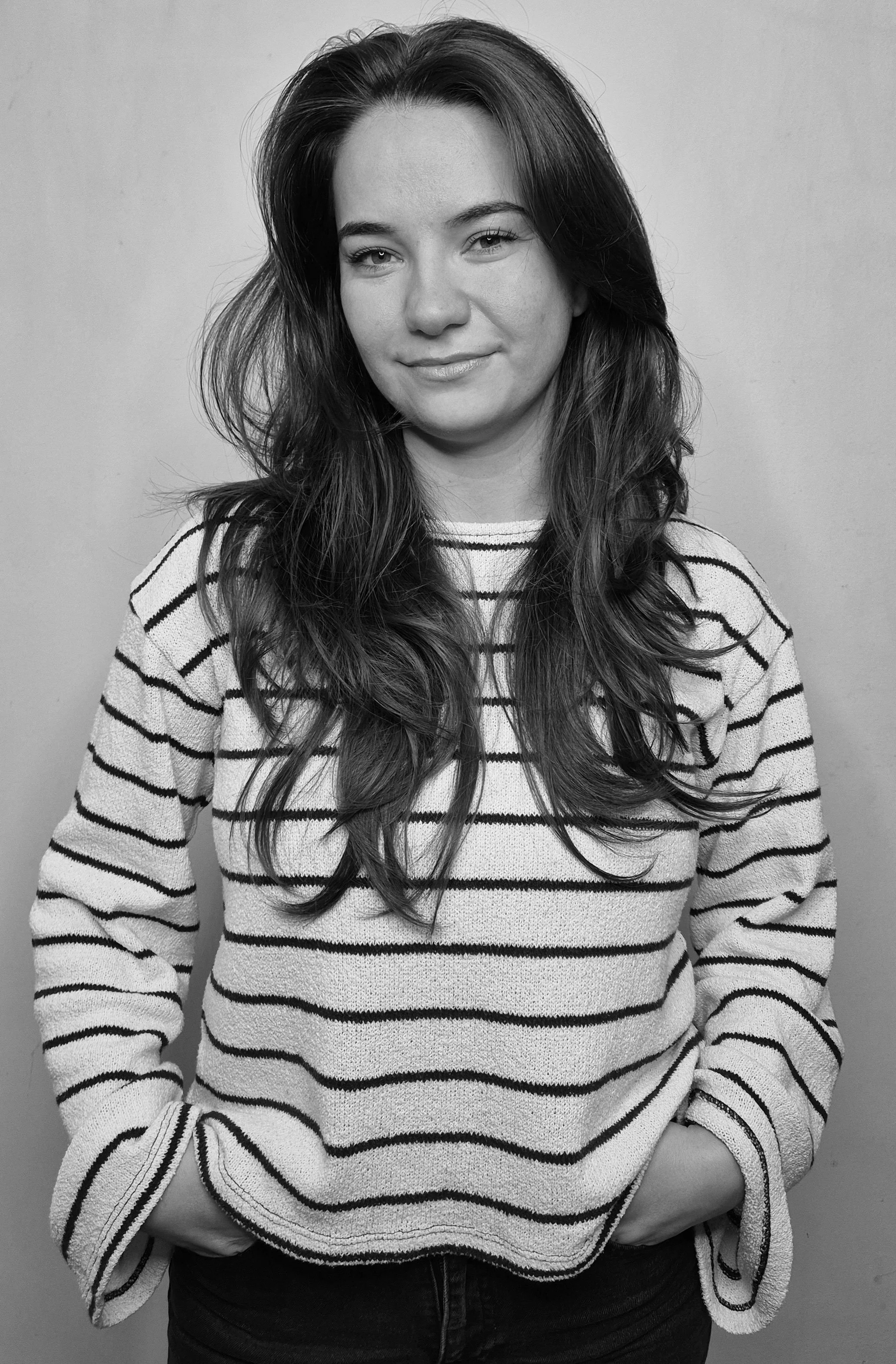 Black and white portrait of a woman with long wavy hair, wearing a black top, standing against a plain wall.
