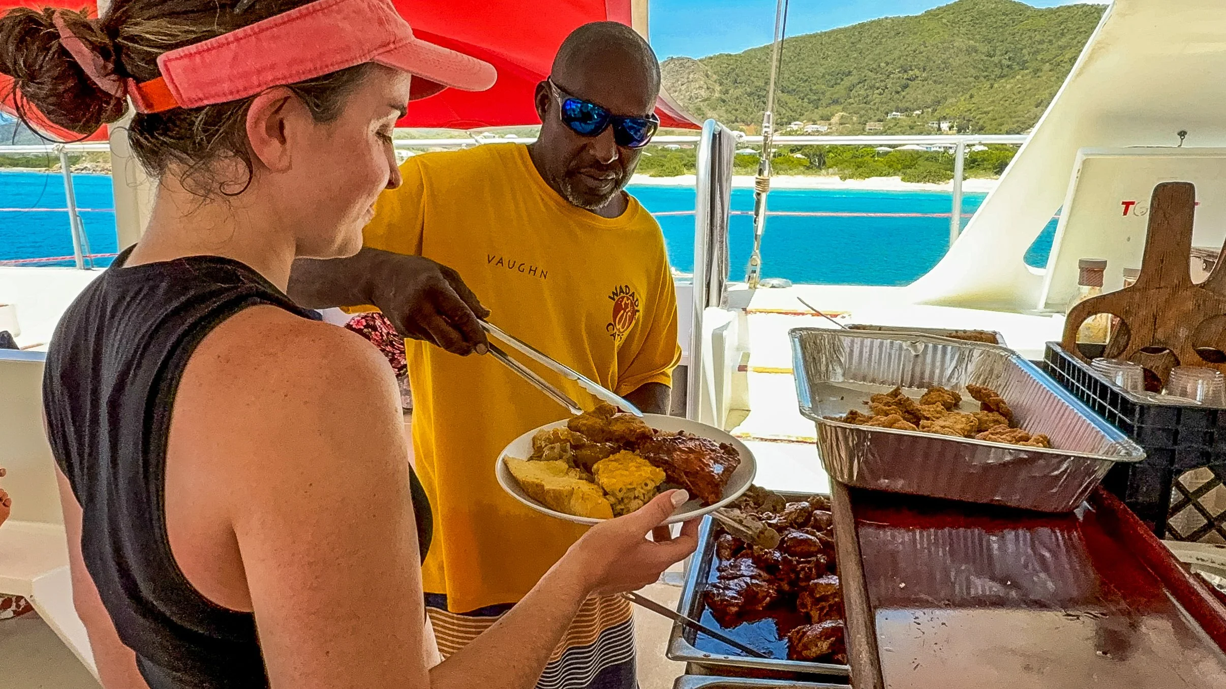 A woman with a pink hat and black tank top is holding a plate of food in front of a man wearing sunglasses and a yellow shirt, serving food at an outdoor buffet on a boat with a tropical ocean and green island in the background.
