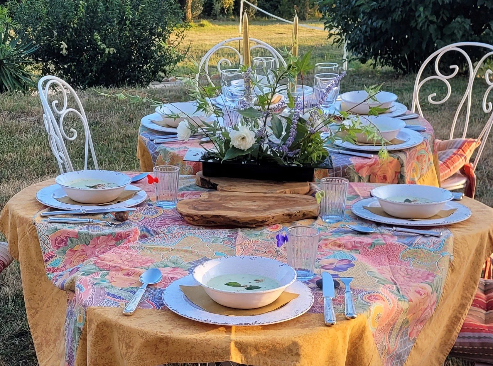 An outdoor dining table set for six with plates, bowls, glasses, silverware, and a flower centerpiece, surrounded by white wrought iron chairs on a grassy yard.