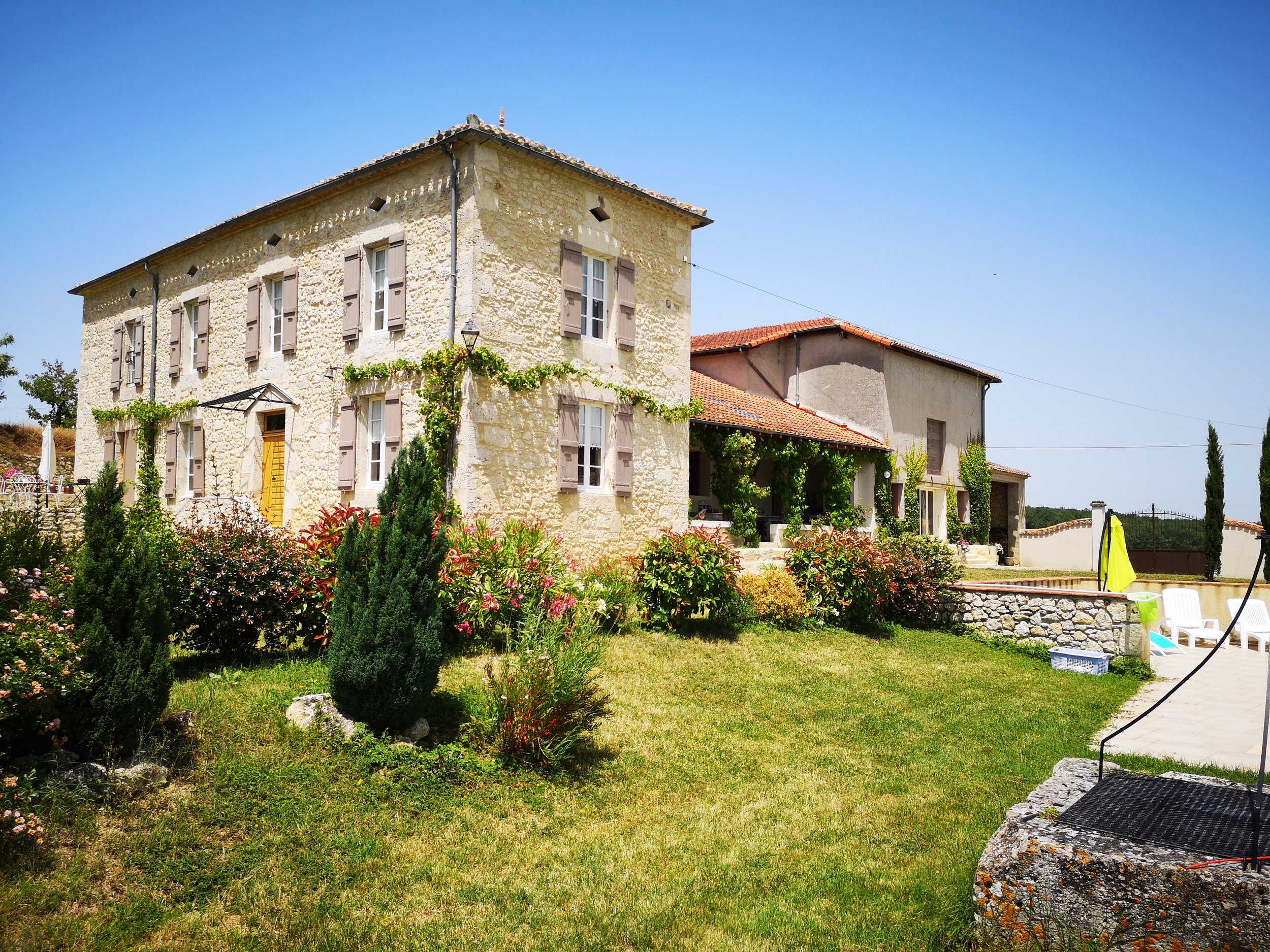 A large stone house with multiple windows, surrounded by a landscaped garden with shrubs and flowers, under a bright blue sky.
