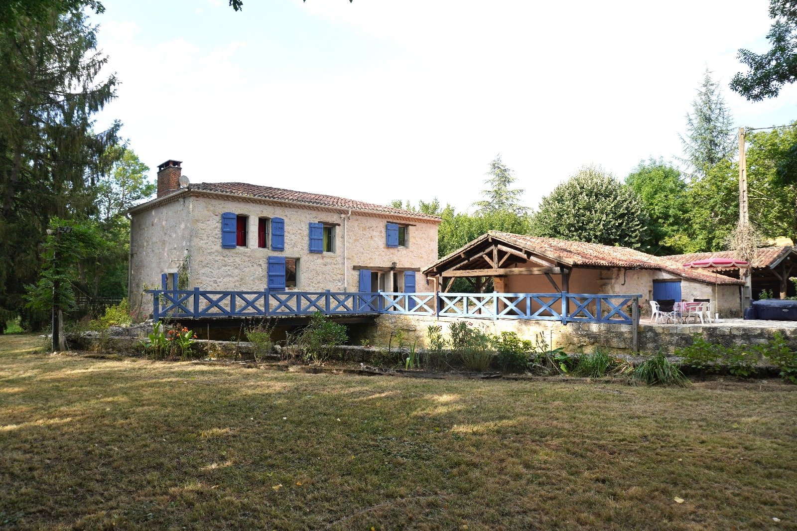A stone house with blue window shutters and a blue railing deck, situated in a lush garden with tall trees in the background and a patio with outdoor furniture.