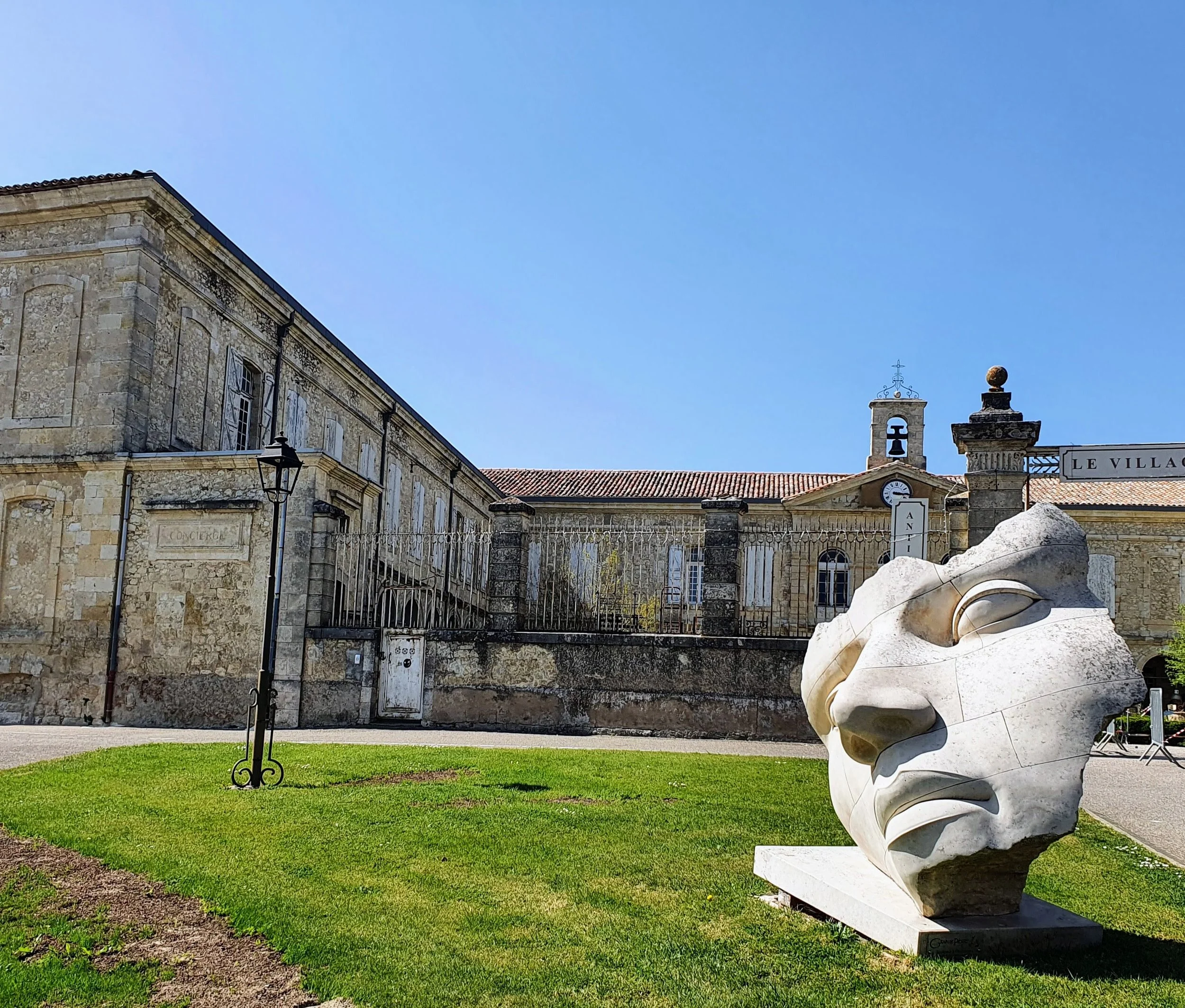 White abstract sculpture of a human face on a pedestal in front of a historic stone building with a small bell tower, set on a grassy area under a clear blue sky.