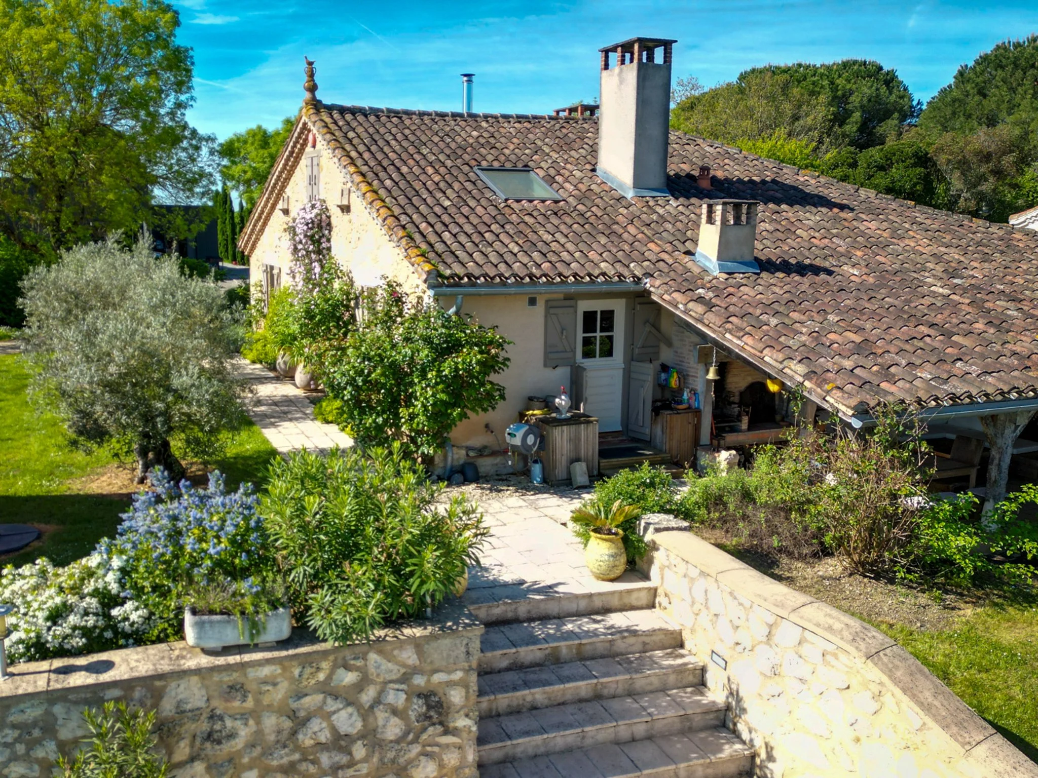 A charming countryside house with a tiled roof, surrounded by lush greenery and flowers. Stone steps lead up to the house, with potted plants along the pathway. The house features closed window shutters, a chimney, and an outdoor seating area under a