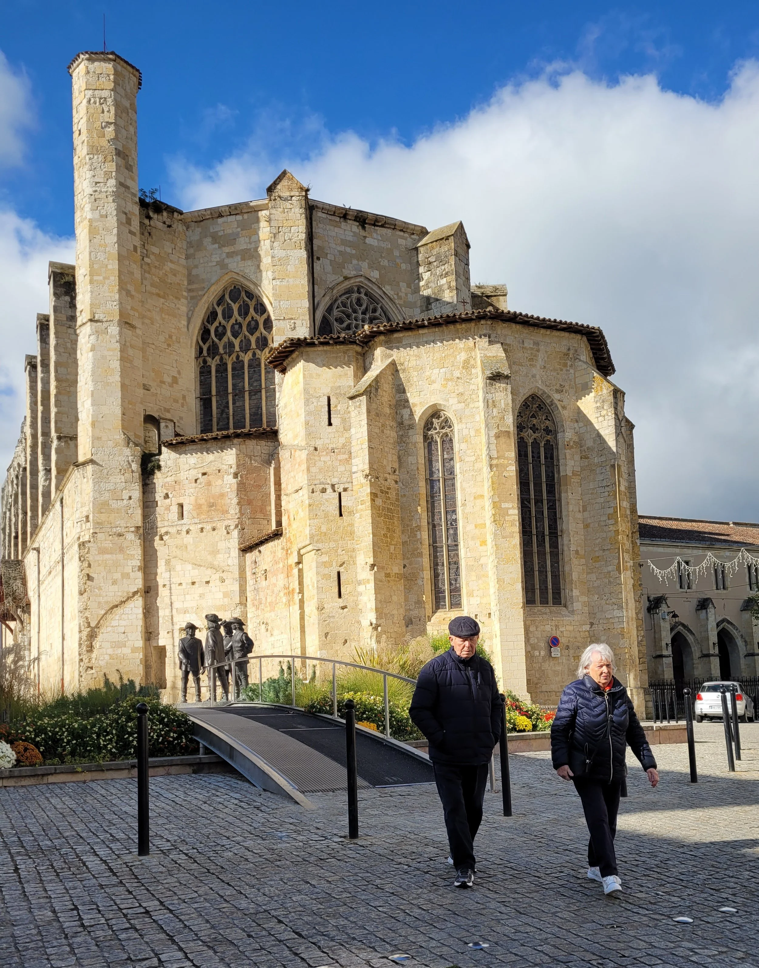 Two elderly people walking in front of a historic stone church with large stained glass windows, set against a partly cloudy sky.