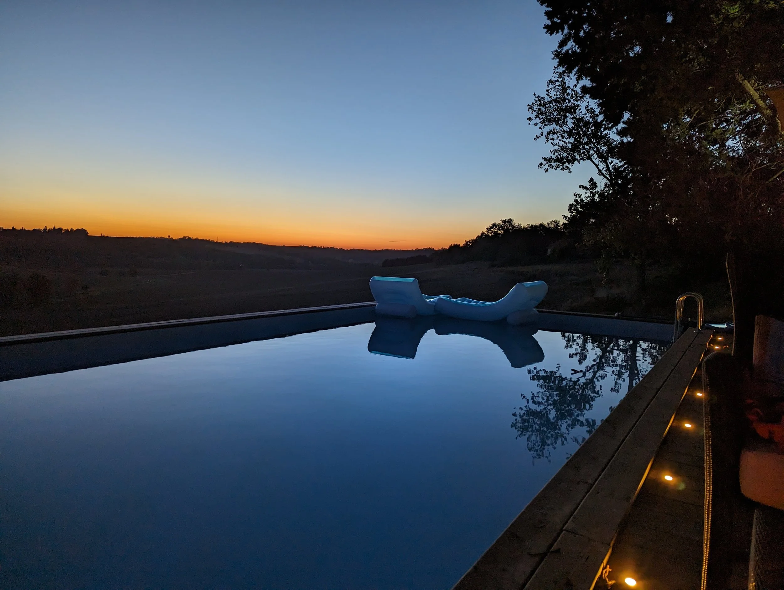 A backyard swimming pool at sunset with an inflatable lounge chair on the pool's edge, surrounded by trees and outdoor string lights.