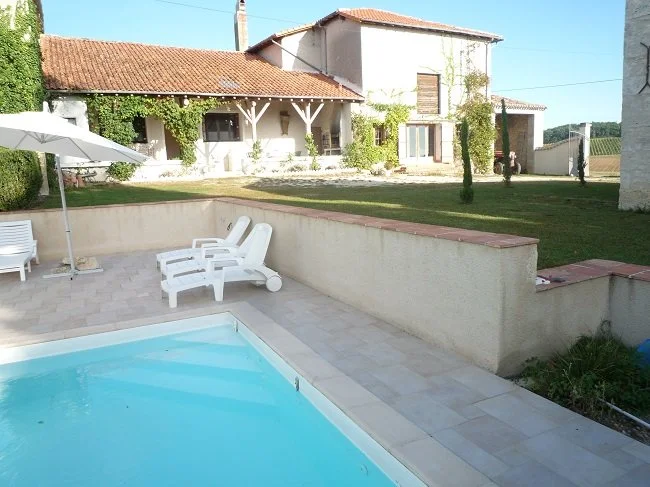 A backyard with a swimming pool, lounge chairs, an umbrella, a grassy area, and a house in the background.