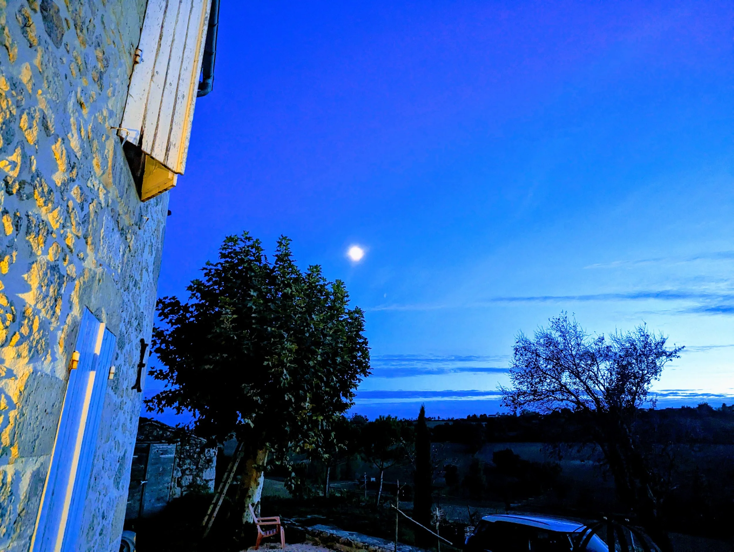 Nighttime scene with a clear sky and visible moon, trees in the yard, a stone house wall on the left, and a parked vehicle in the background.