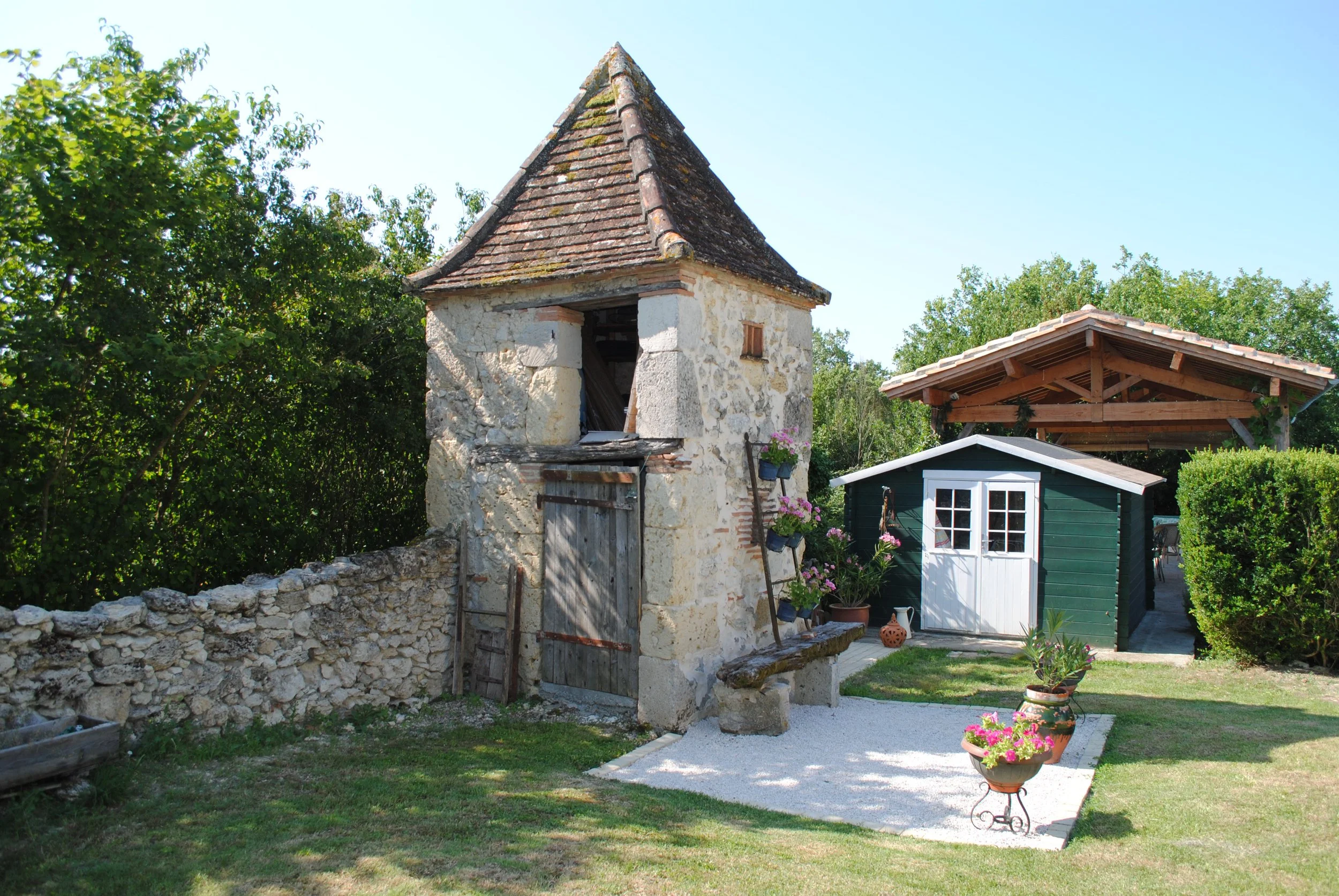 A small stone tower with a conical tiled roof, a wooden door, and a small window on the side surrounded by greenery and a garden with potted plants, a green shed with a white door, and a wooden open structure in the background under a clear blue sky.