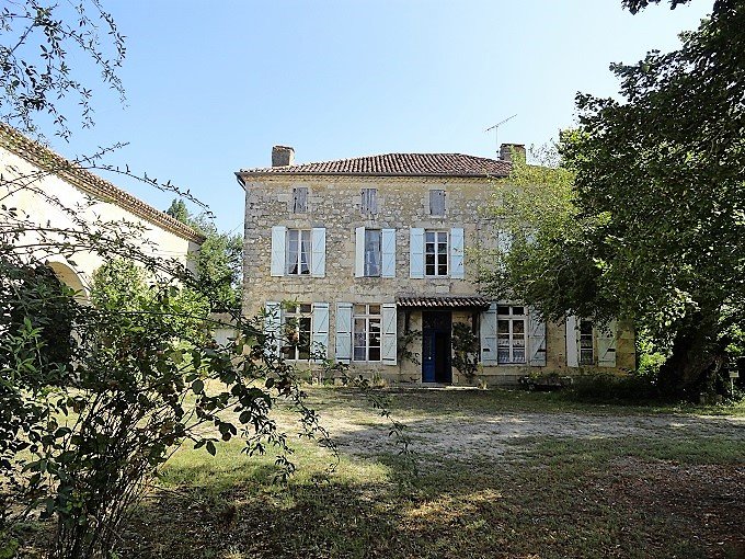 An old stone house with multiple windows and blue shutters, surrounded by trees and bushes, under a clear blue sky.
