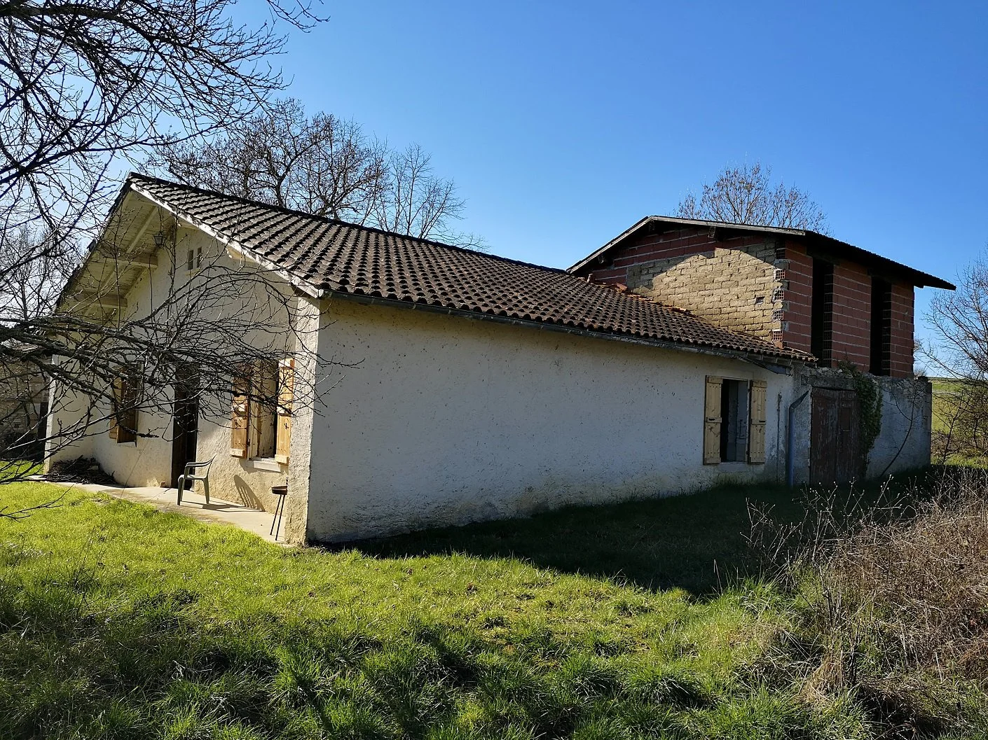 A house with a tiled roof, white exterior walls, and wooden shutters on the windows. There is a small porch with a chair and a table outside. The background features leafless trees and a clear blue sky.
