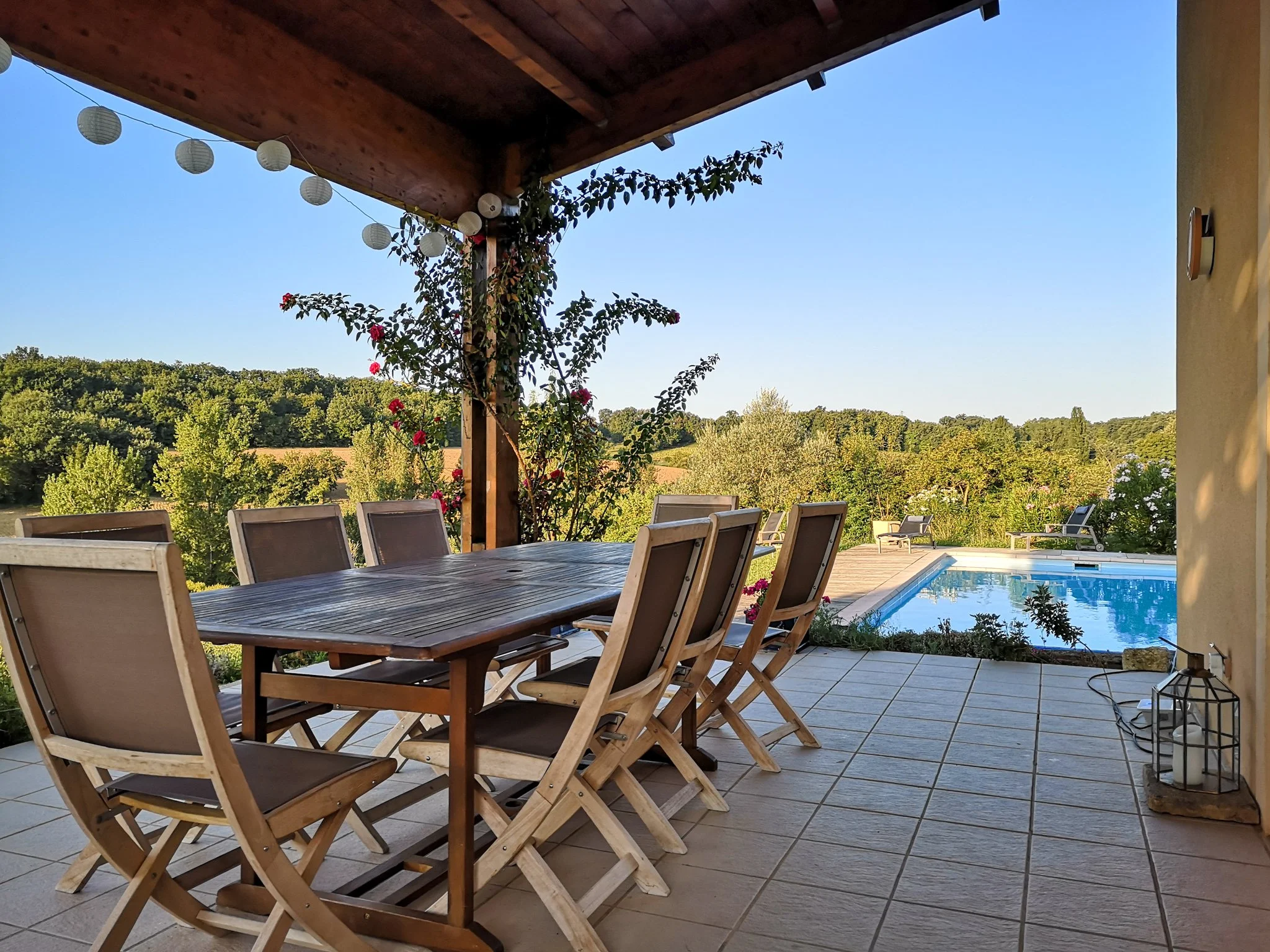 Outdoor patio with a wooden dining table and chairs, overlooking a swimming pool and a lush green landscape under a clear blue sky, decorated with string lights and potted plants.