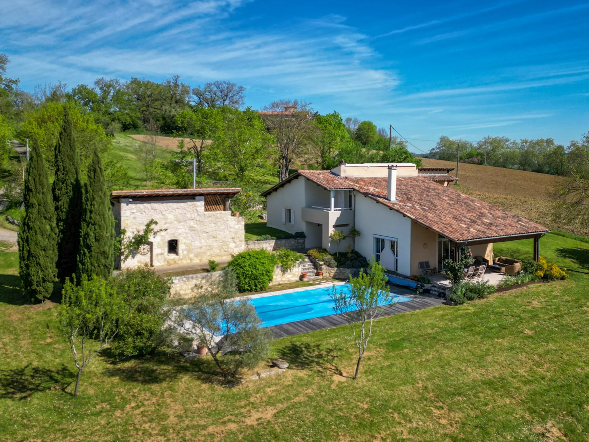 A house with a swimming pool in a green yard, surrounded by trees and hills, under a blue sky.