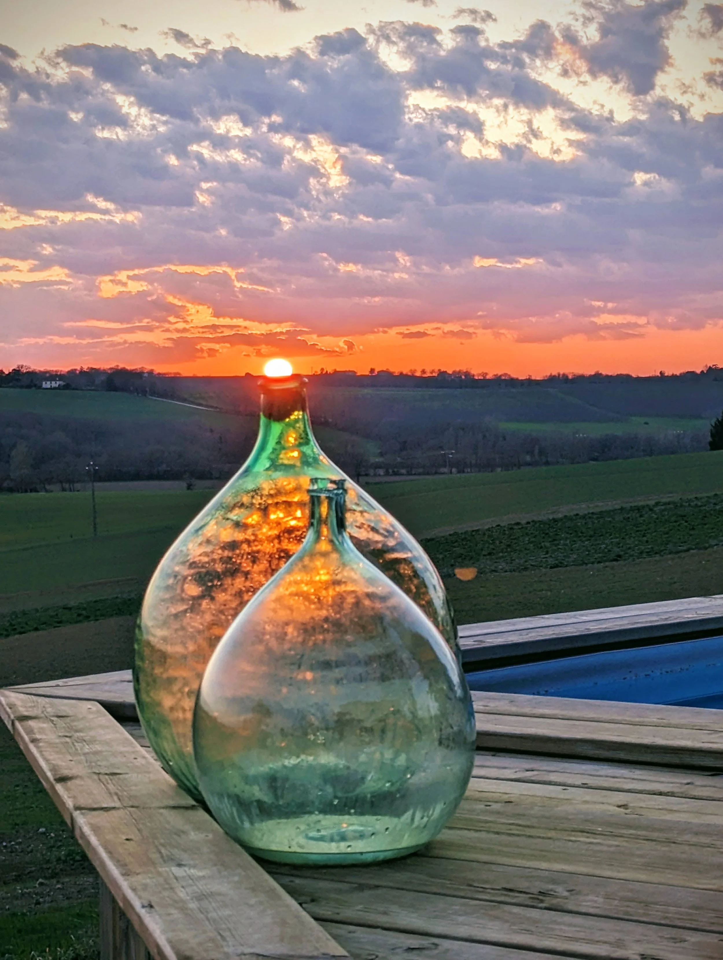 A scenic sunset landscape with colorful clouds over green fields, seen through the reflection in a glass flask on a wooden table.