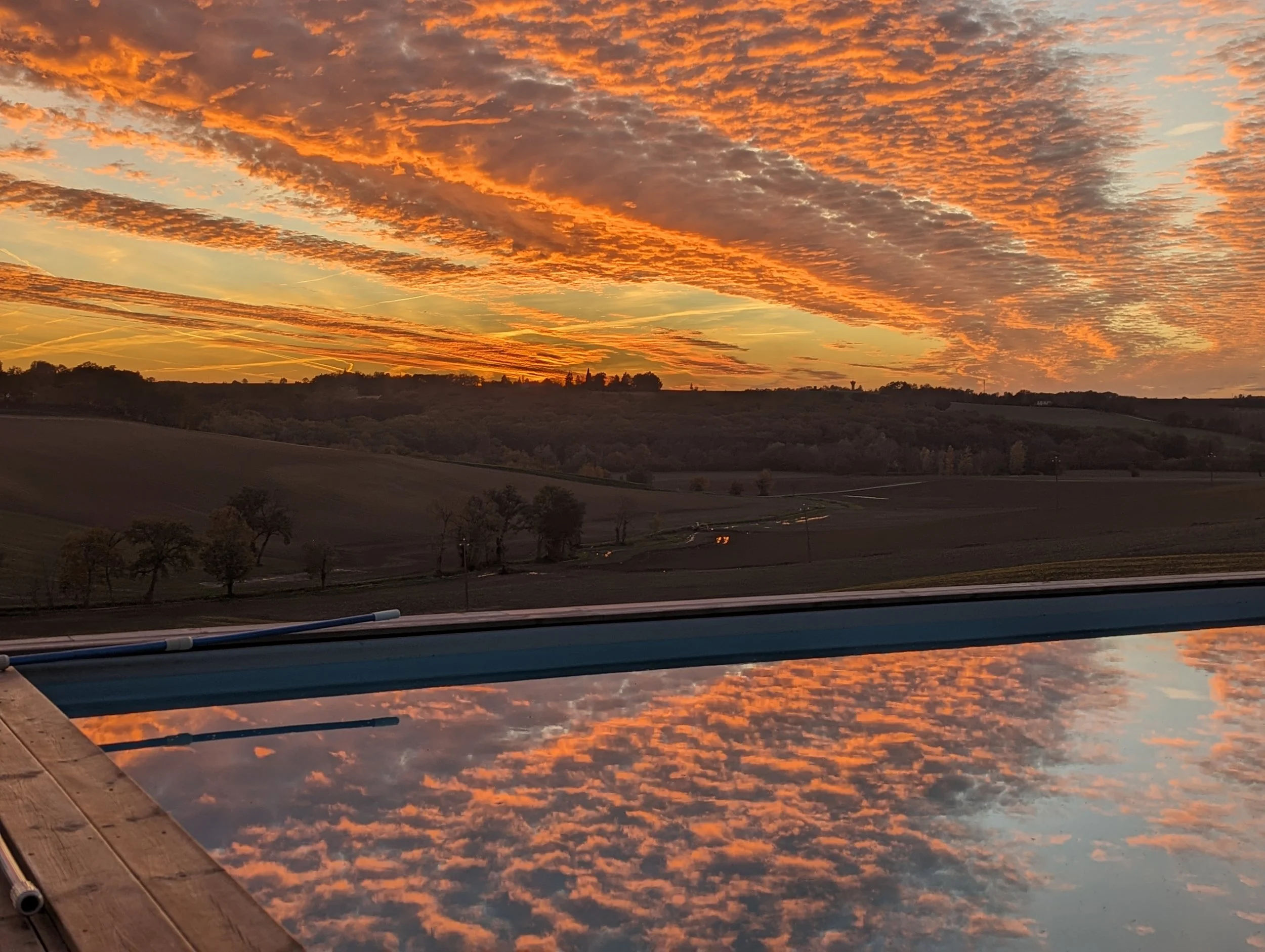 Sunset over a rural landscape with scattered trees, reflected in a pool of water in the foreground, creating a mirror image of the colorful sky.