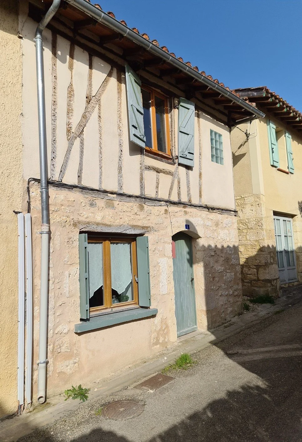 A stone and stucco house with green shutters, a green door, and a small window, on a narrow street with a drain grate and a bit of grass, under a clear blue sky.