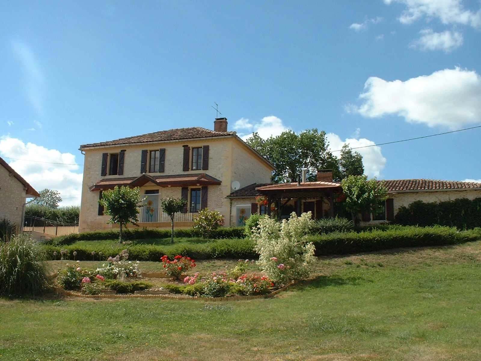 A large two-story house with beige stone walls, brown shutters, and a brown tiled roof, surrounded by a green lawn and colorful flower beds, under a blue sky with white clouds.