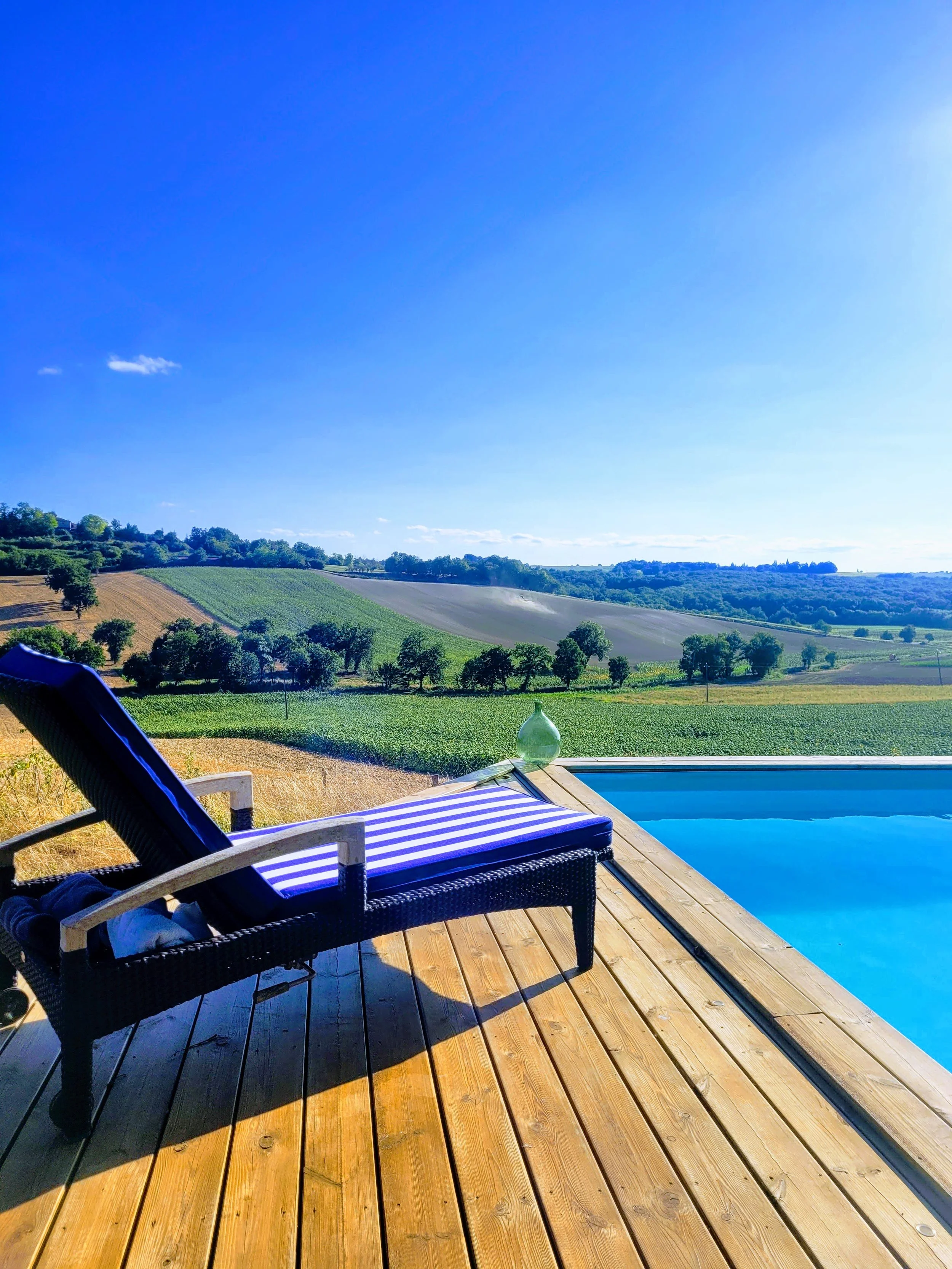 A poolside scene with a lounge chair and a glass bottle on a wooden deck overlooking green rolling hills under a bright blue sky.