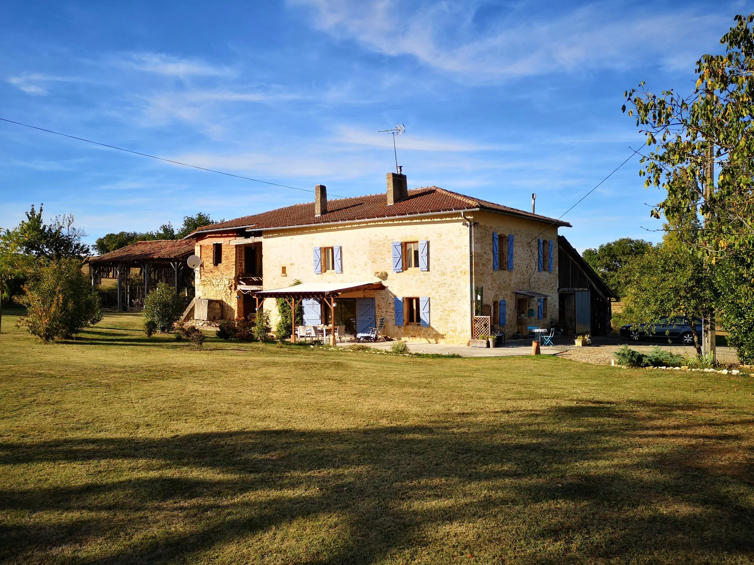 A rustic two-story house with blue shutters and a tiled roof, surrounded by a large lawn with trees, and a clear blue sky overhead.