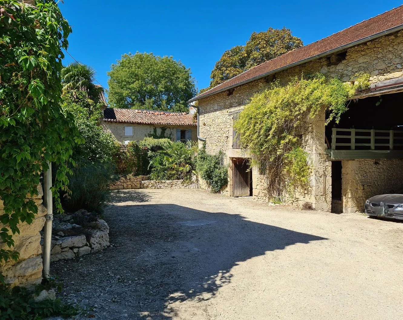 A stone farmhouse with a tiled roof, surrounded by greenery, under a clear blue sky. There is a small courtyard with gravel and a car parked to the right.