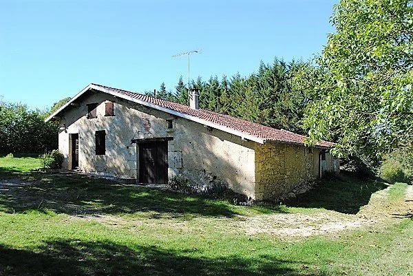 An old, rustic house with a sloped roof surrounded by grass and trees under a clear blue sky.