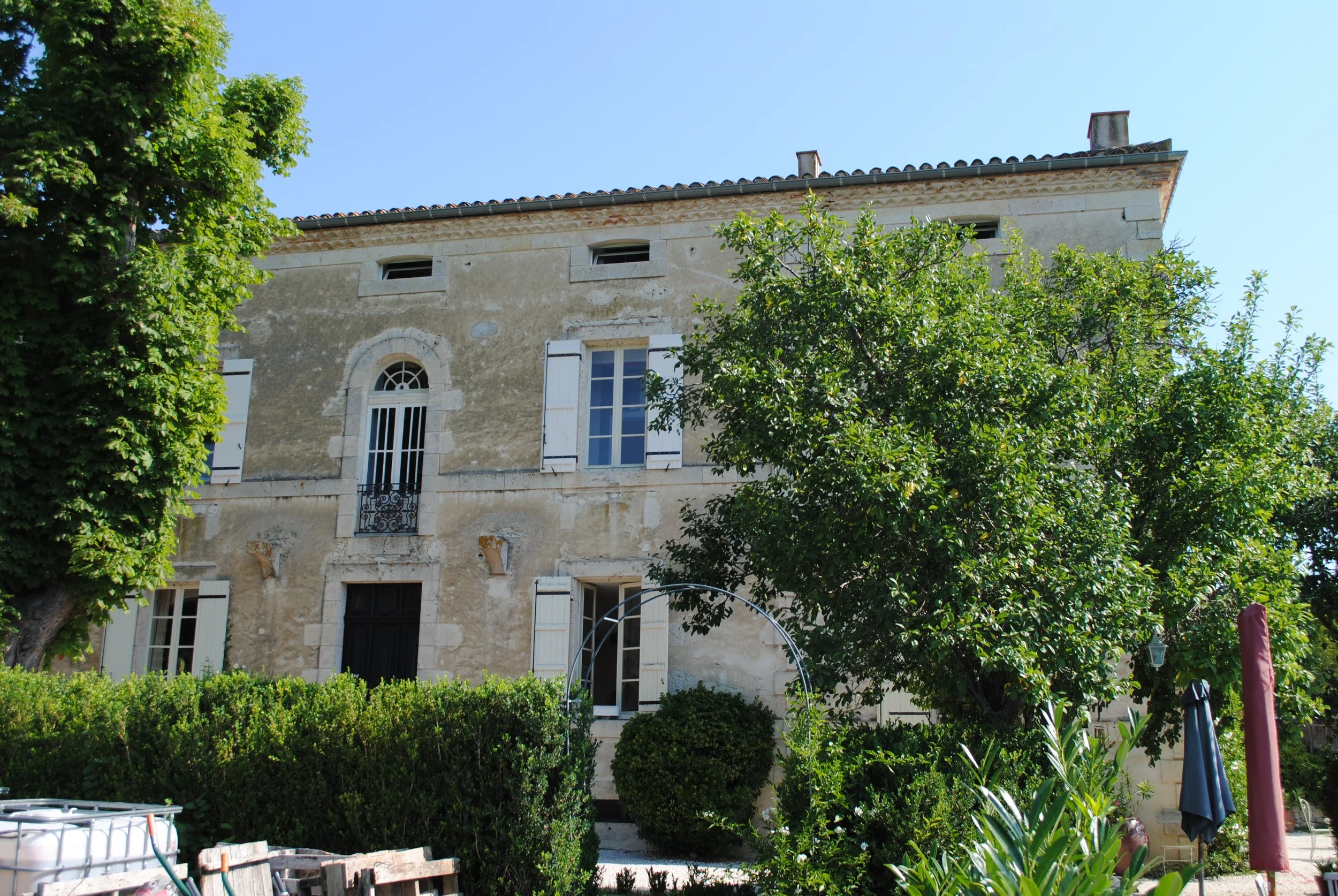 A large, old stone building with white window shutters, surrounded by lush green trees and bushes, under a clear blue sky.