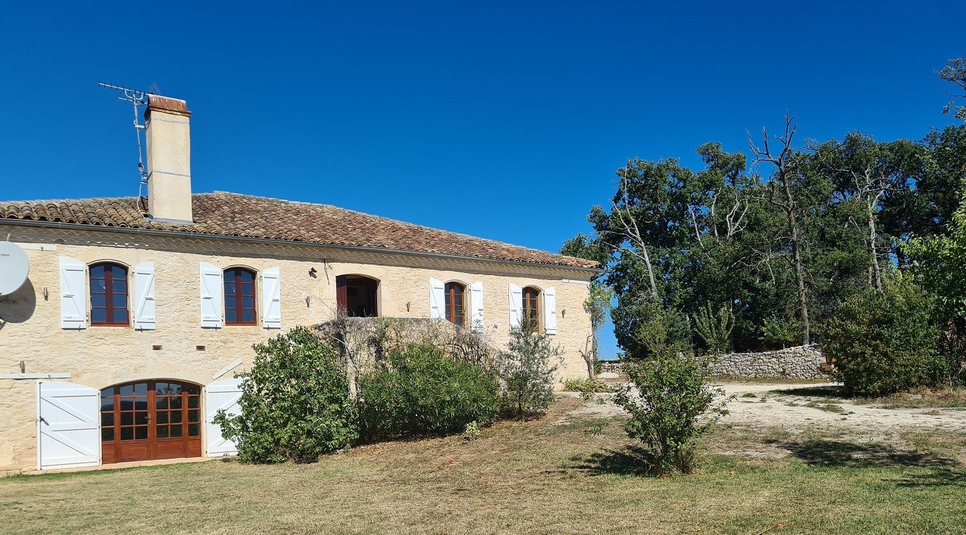 Stone house with white shutters and brown wooden doors, surrounded by trees and shrubs under a clear blue sky.