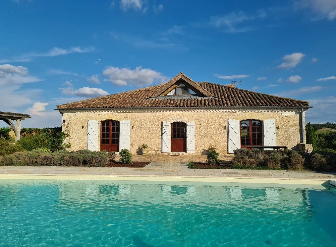 Stone house with white shutters, red doors, and a tiled roof in a rural setting, with a swimming pool in the foreground and a blue sky with some clouds in the background.