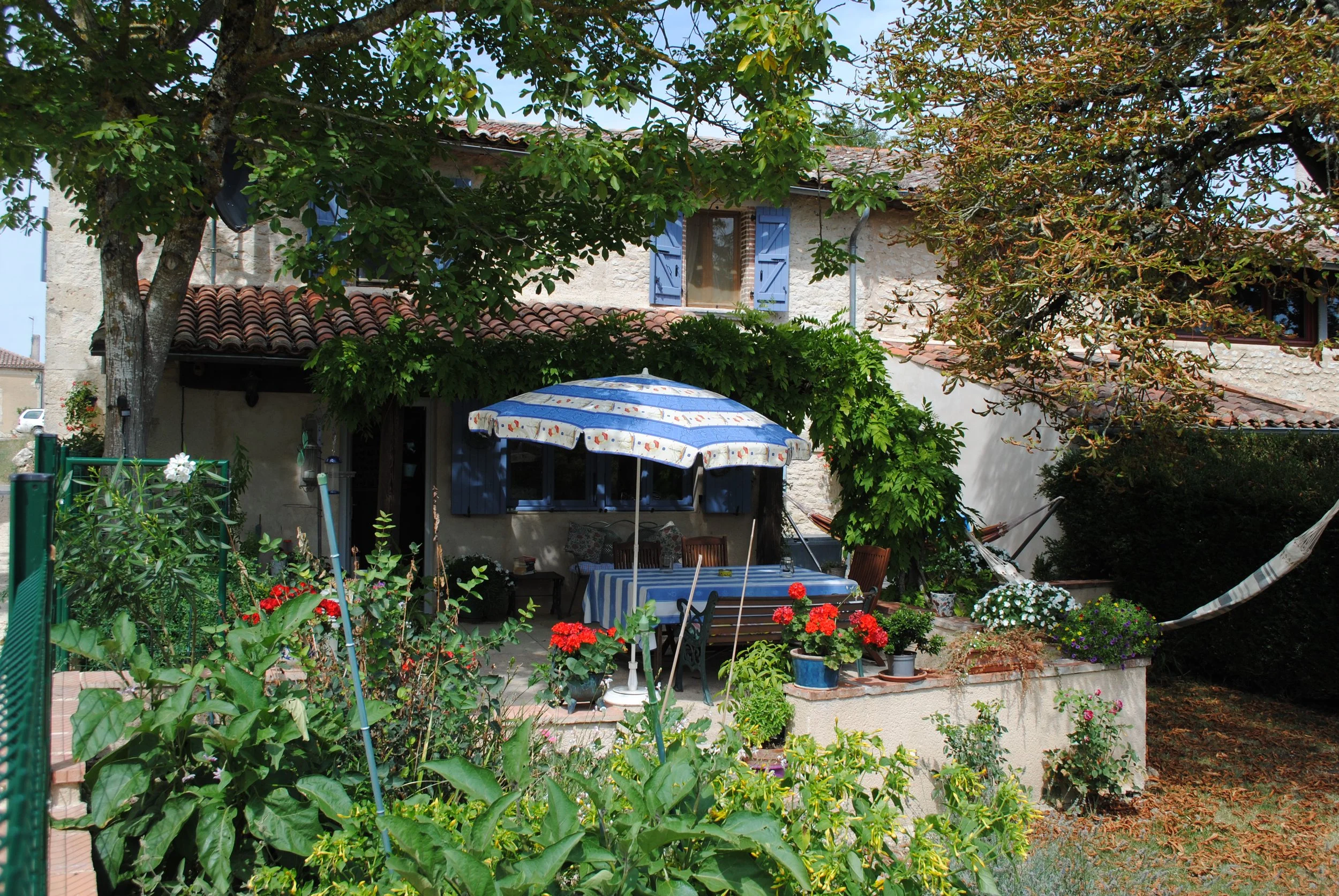 A cozy outdoor patio garden with a table and chairs under a large umbrella, surrounded by potted plants and flowers, with a house in the background.