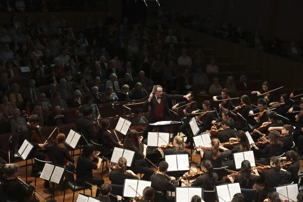 An orchestra performs on stage with a conductor in front, playing string instruments while an audience watches in a concert hall.