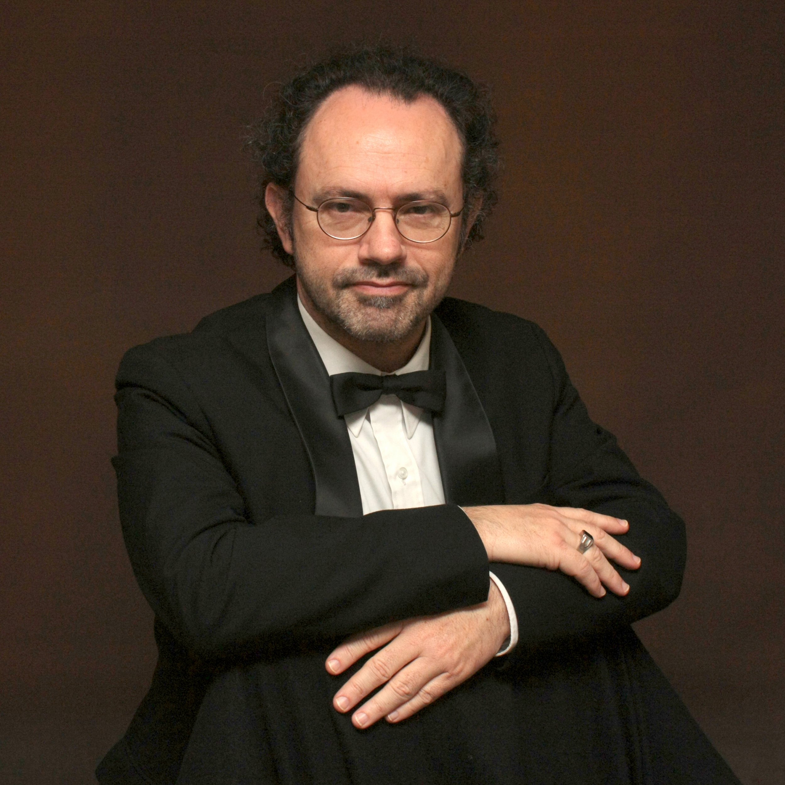 A man with curly hair, glasses, and a beard, wearing a tuxedo with a bow tie, posing with arms crossed against a brown background.