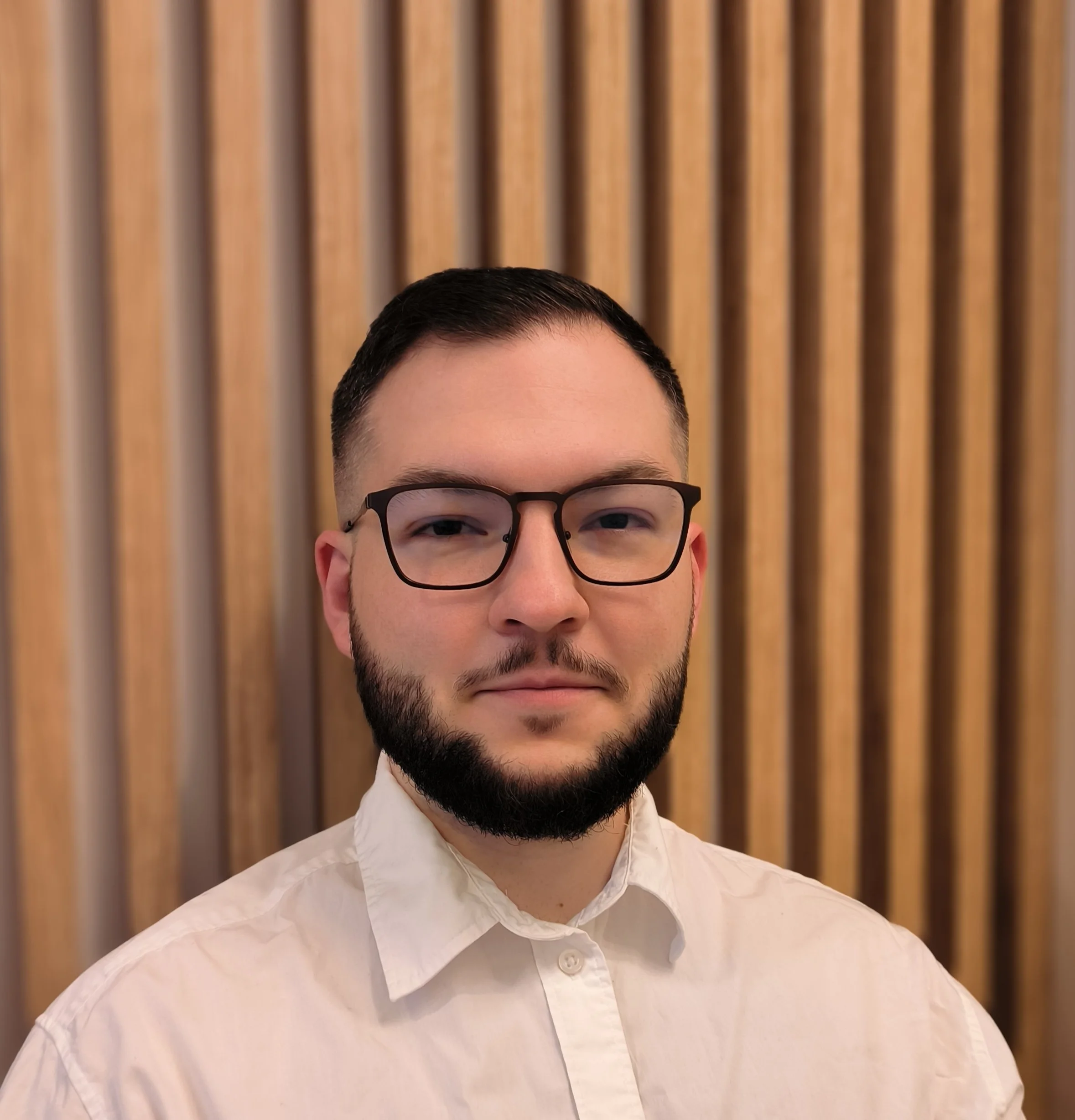 A man with short dark hair, glasses, and a beard wearing a white button-up shirt, standing in front of a wooden slat background.