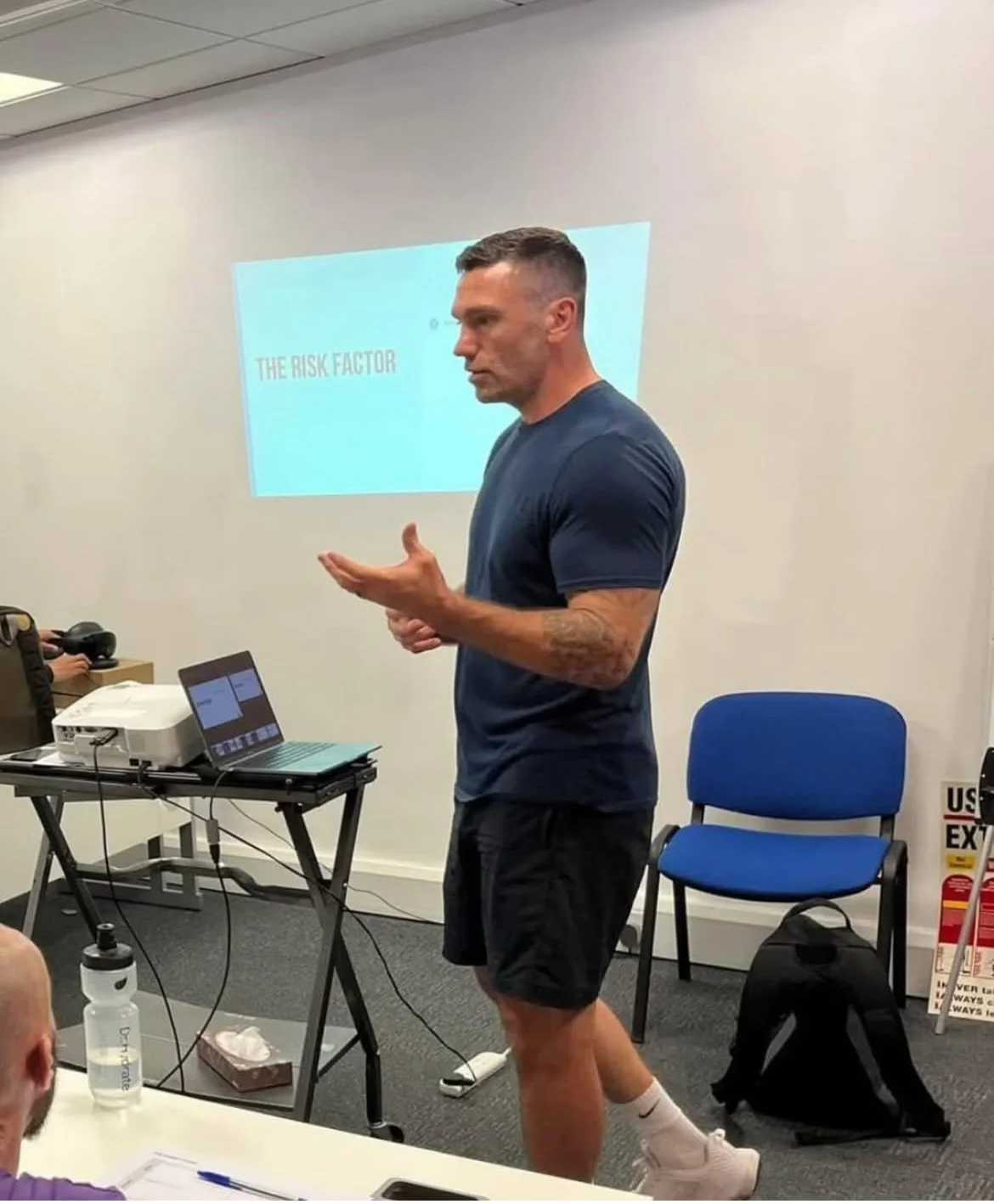 Man giving presentation in a classroom or training room, with a slide titled 'The Risk Factor' projected on the wall behind him.