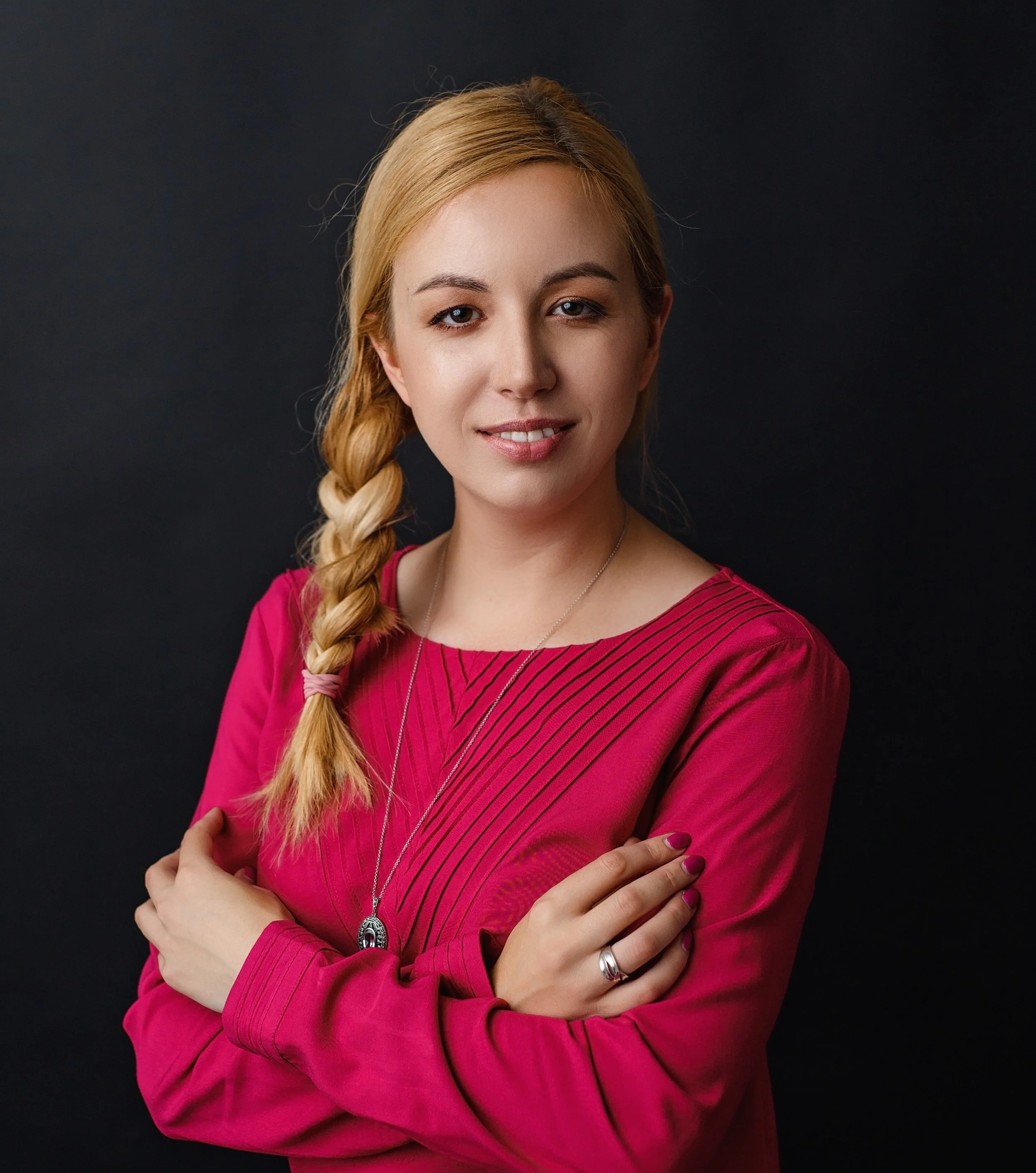 Alicia Skuza with blonde hair styled in a side braid, wearing a pink dress, standing against a dark background.