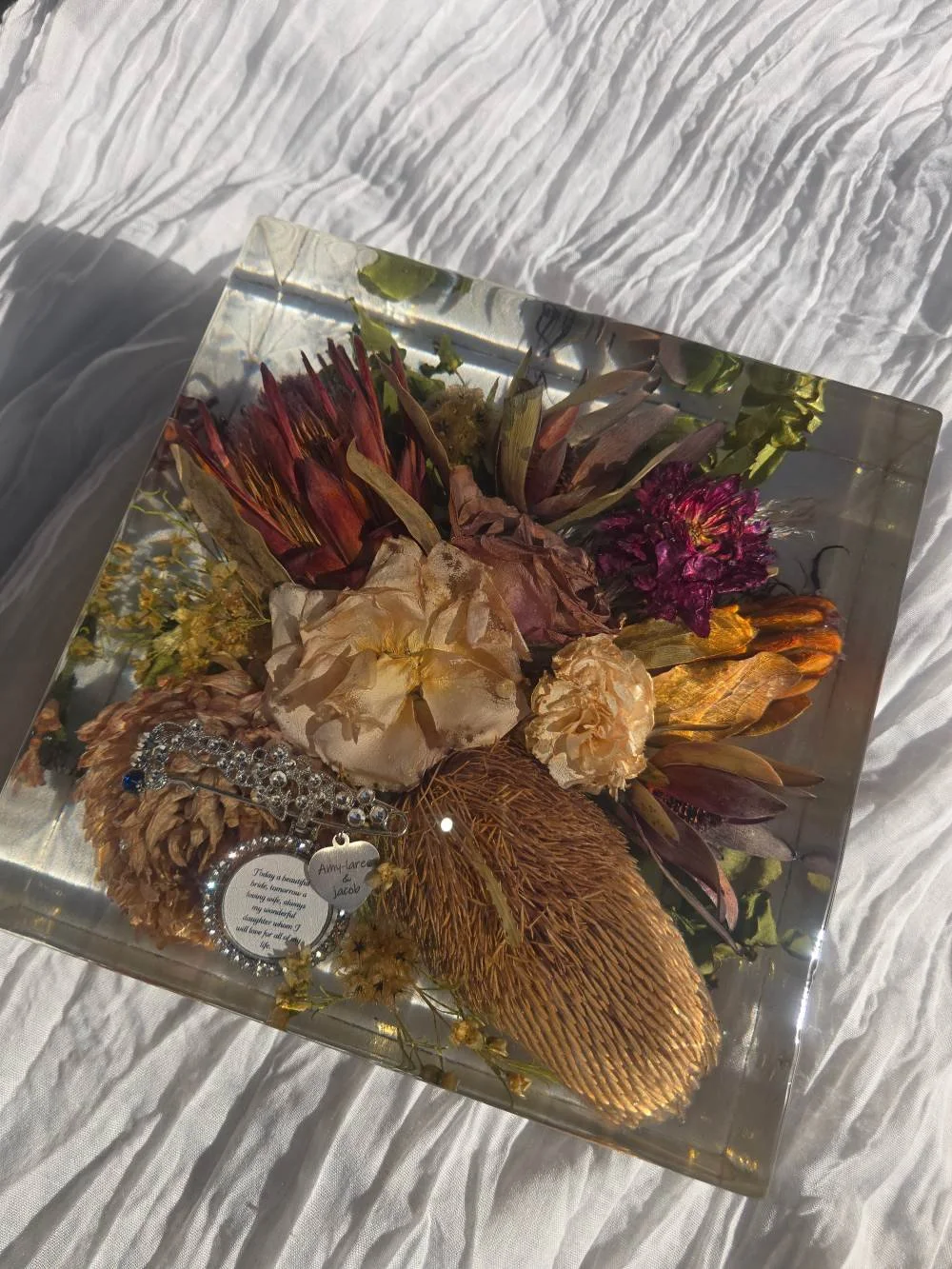 A decorative display of dried flowers, a bracelet, and a small tag inside a glass box, placed on a white textured surface.