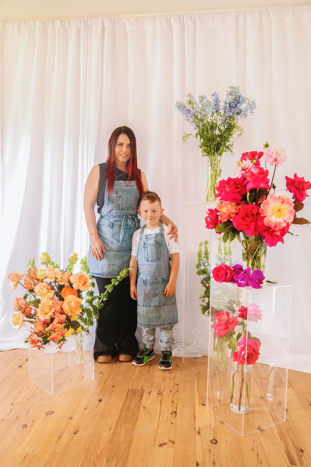 A woman and a young boy standing together in front of a white curtain, surrounded by large colorful floral arrangements in glass vases, including pink, red, purple, and peach flowers.