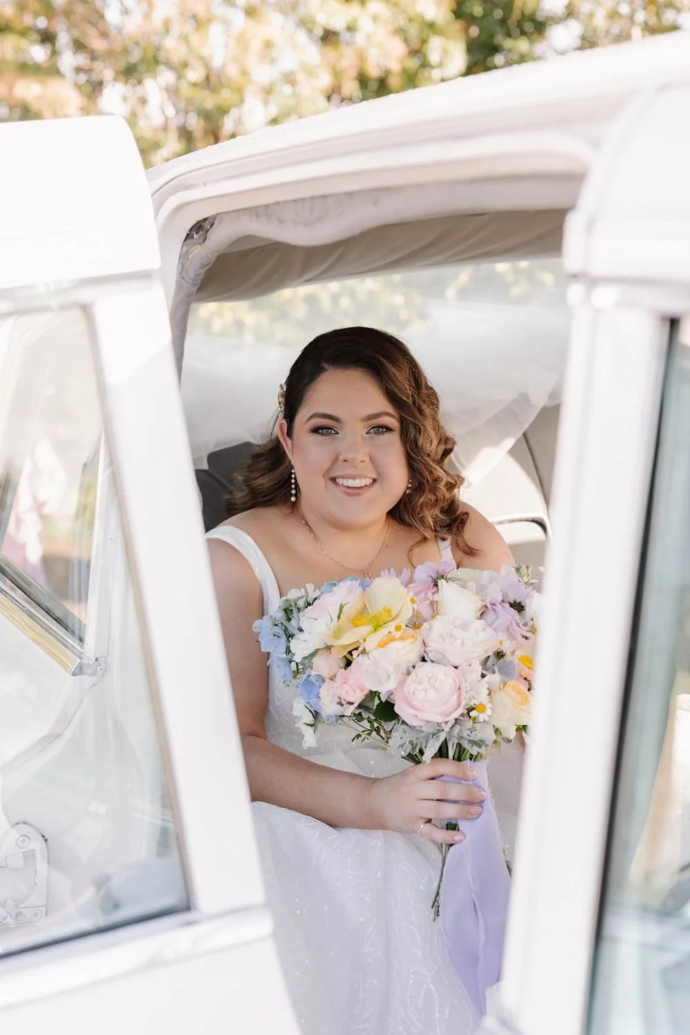 A bride sitting inside a white vintage car, holding a bouquet of pink and white flowers, smiling at the camera.