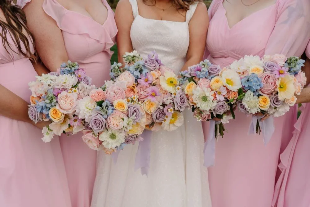 A bride in a white wedding dress is standing with bridesmaids dressed in light pink dresses, all holding large bouquets of pastel-colored flowers including roses, peonies, and other blooms.