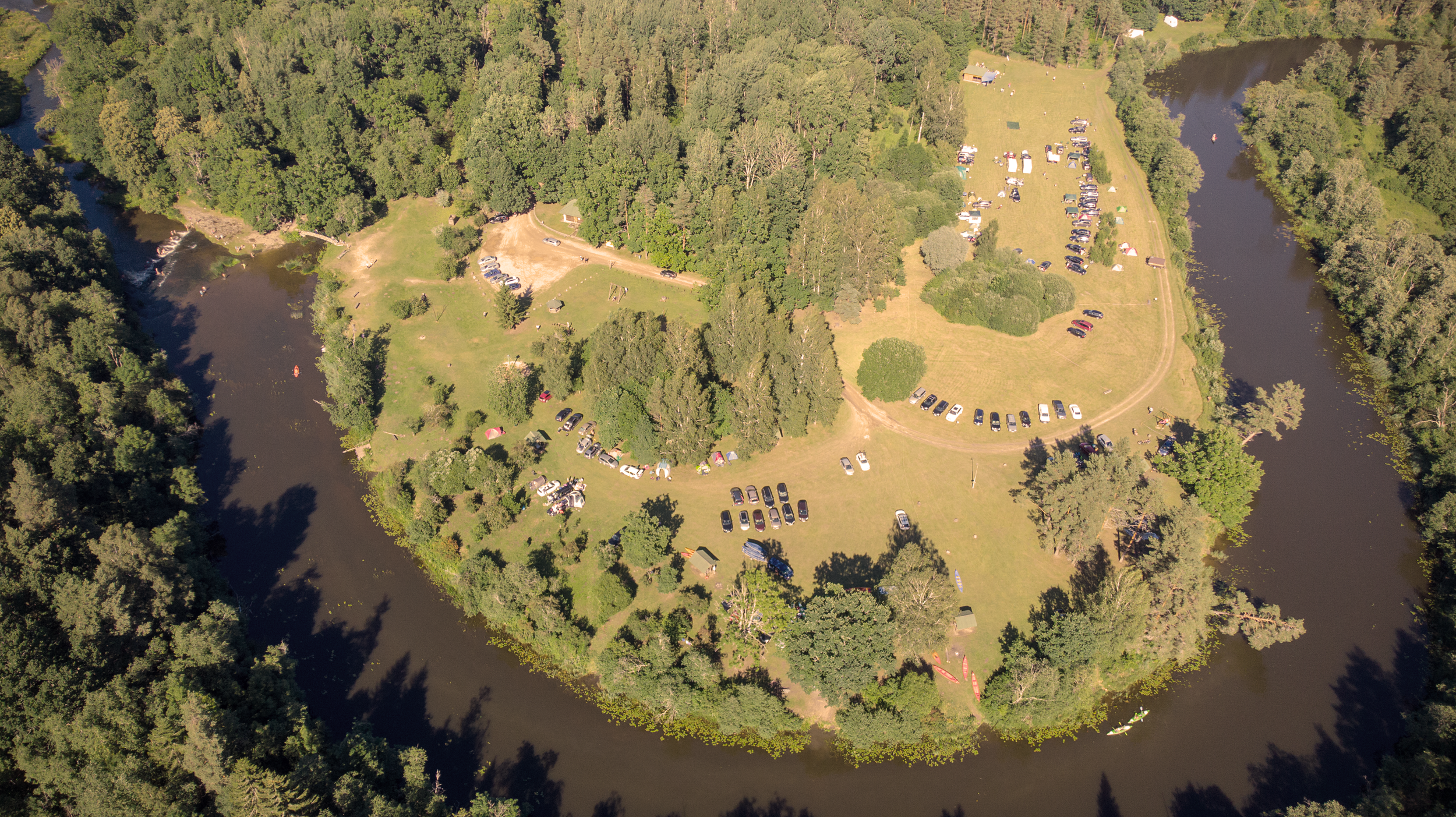 An aerial view of a park surrounded by a river, with parking lots, tents, and a small building visible within the grassy area.