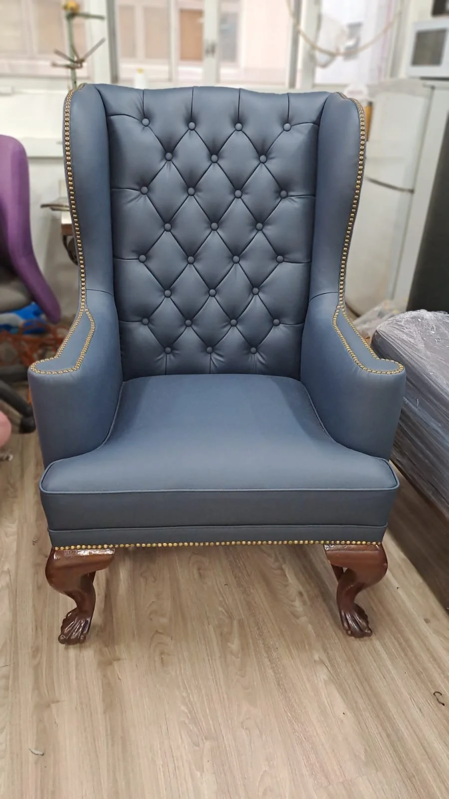 A blue high-back armchair with tufted upholstery, nailhead trim detailing, and wooden carved legs, sitting on a wooden floor in a store or furniture showroom.