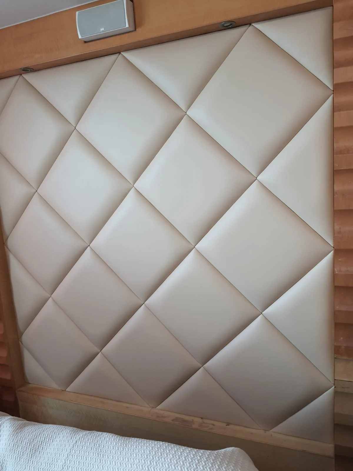 Close-up of a padded headboard with a diamond pattern, in a beige or cream color, attached to a wooden bed frame with a wood-paneled wall in the background.