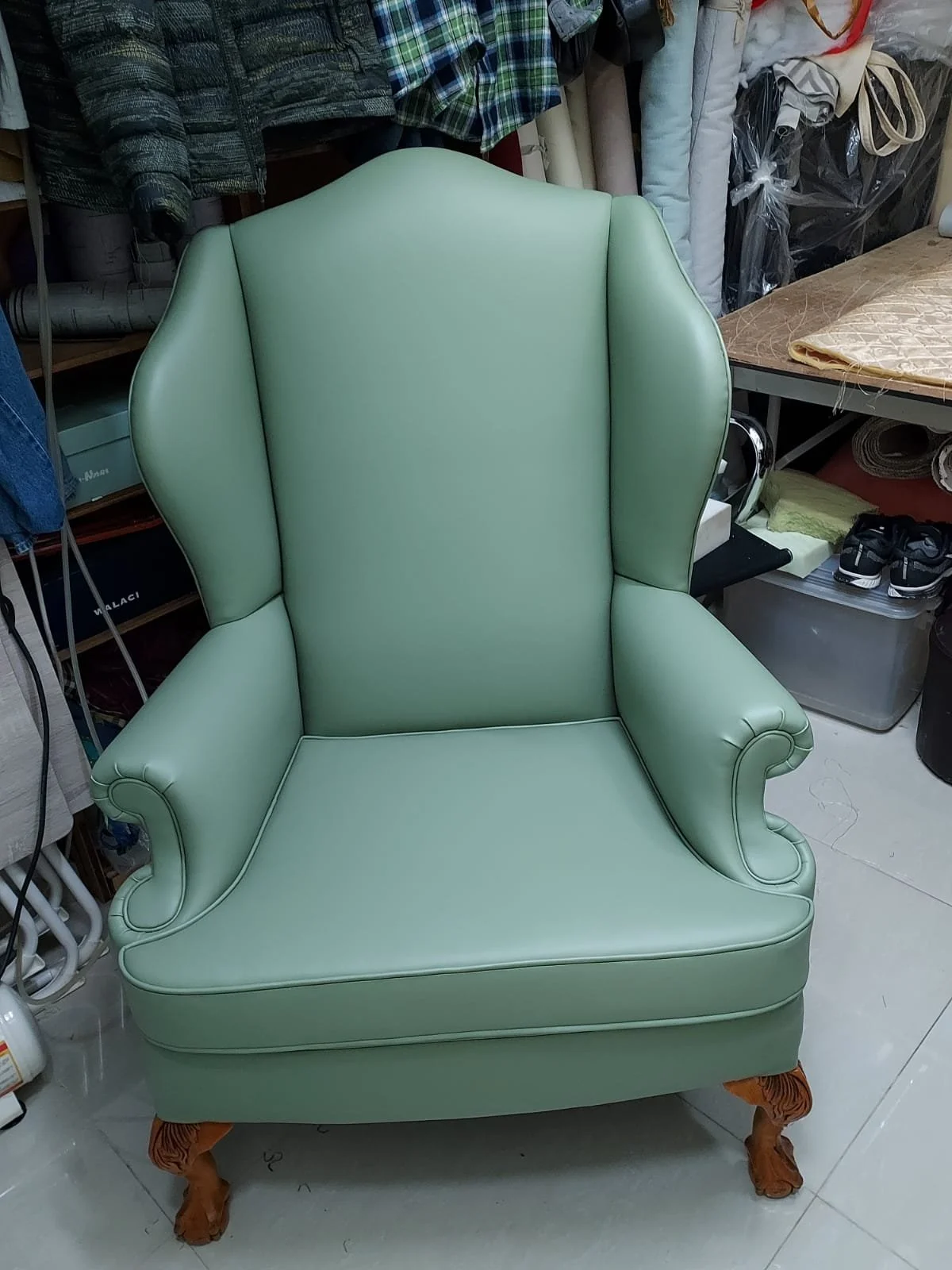 A vintage green high-backed armchair with curved armrests and wooden carved legs, placed in a cluttered room with fabric and other miscellaneous items in the background.