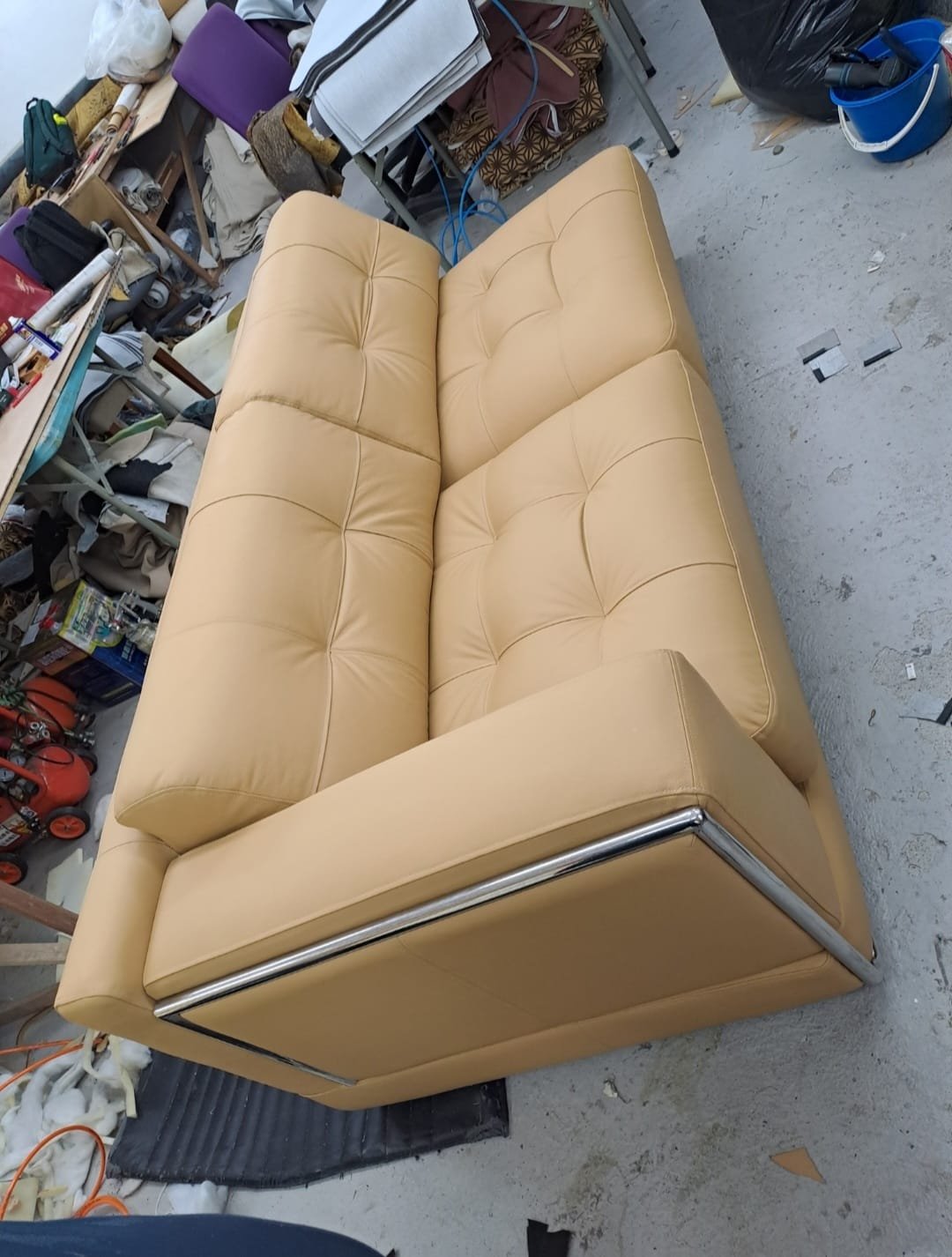 A mustard yellow tufted sofa with metal legs, placed in a cluttered garage or storage space.